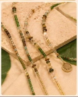 Multiple beaded necklaces with green, gold, and earth-tone beads, some featuring small charms, displayed on a beige surface with green leaves.
