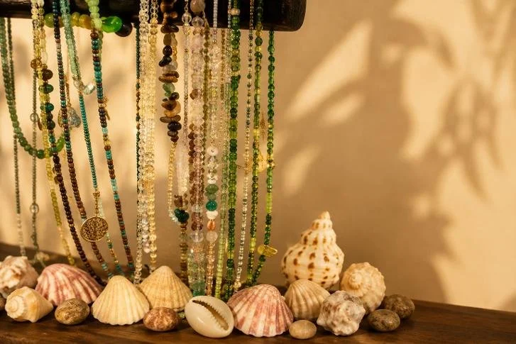 Display of beaded necklaces hanging above seashells and small stones on a wooden surface.