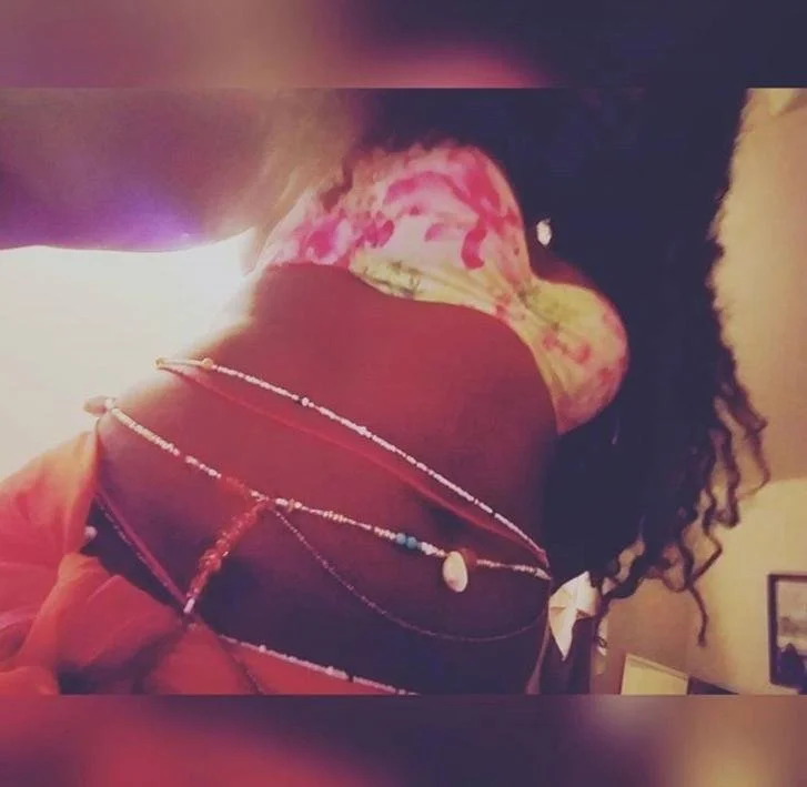 A person with curly hair wearing a black shirt with layered necklaces, including one with a large pendant, and a pink painted chest, taking a photo from below.