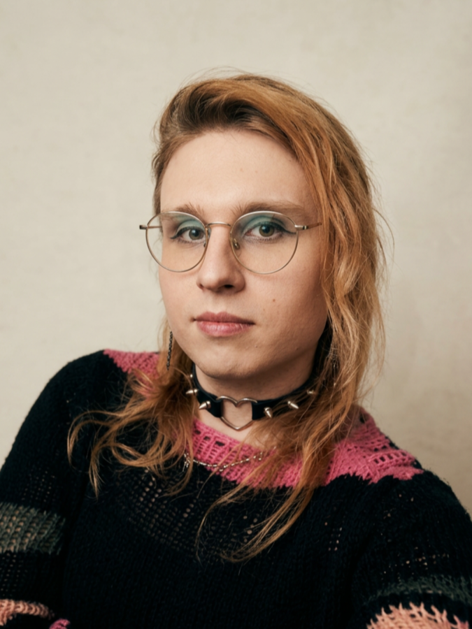 A woman with red hair, wearing glasses with a thin metal frame, a black choker necklace with a heart-shaped ring, and a black sweater with pink and green accents, taking a selfie against a plain background.