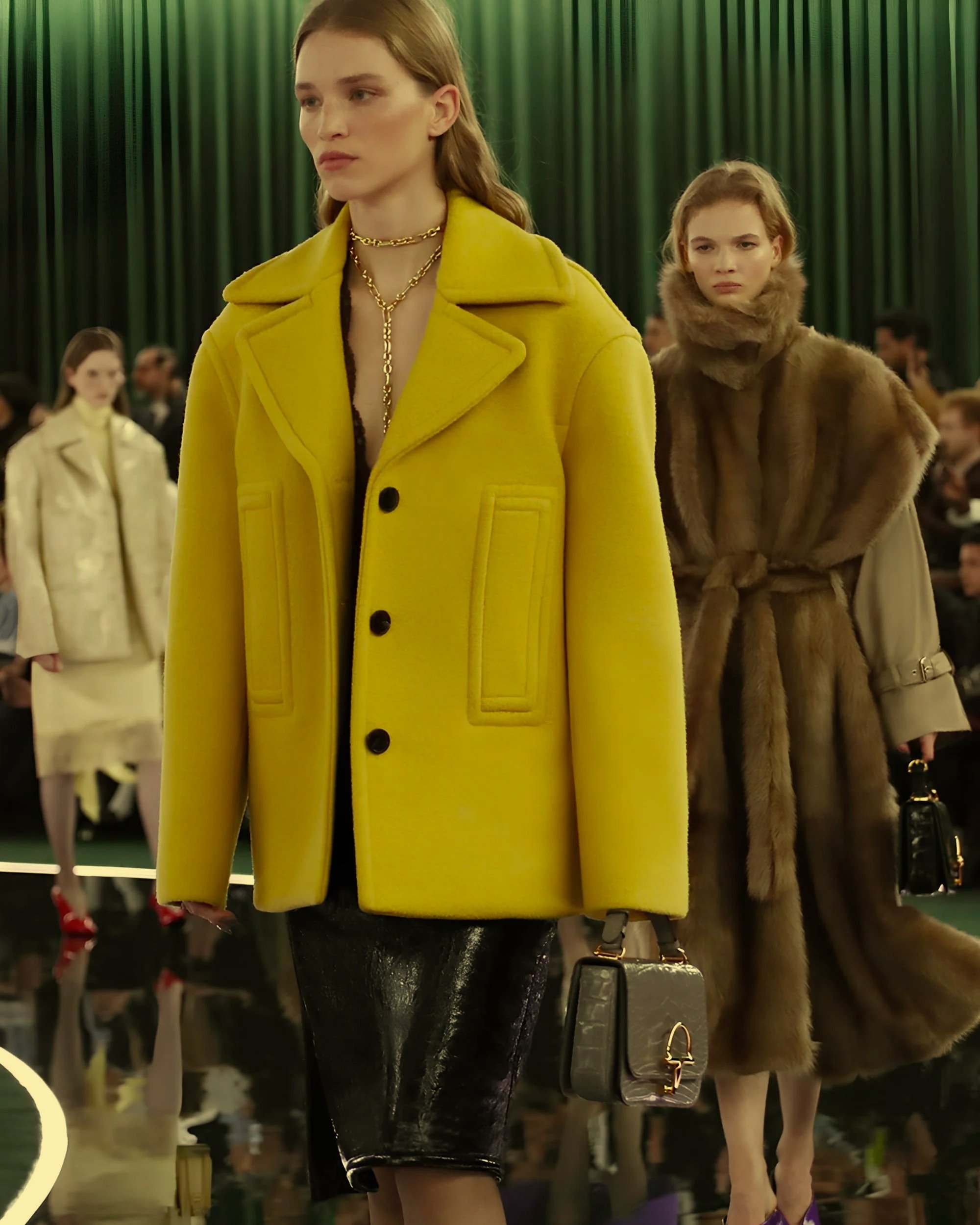 Fashion models walking on runway at fashion show, woman in yellow coat and leather skirt, woman in fur coat and purple shoes, audience in background.