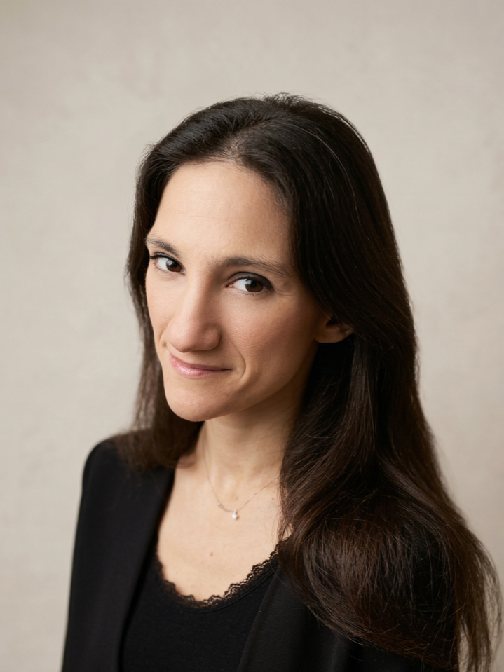 A woman with long dark hair, wearing a black top and blazer, looking at the camera with a slight smile against a plain light background.