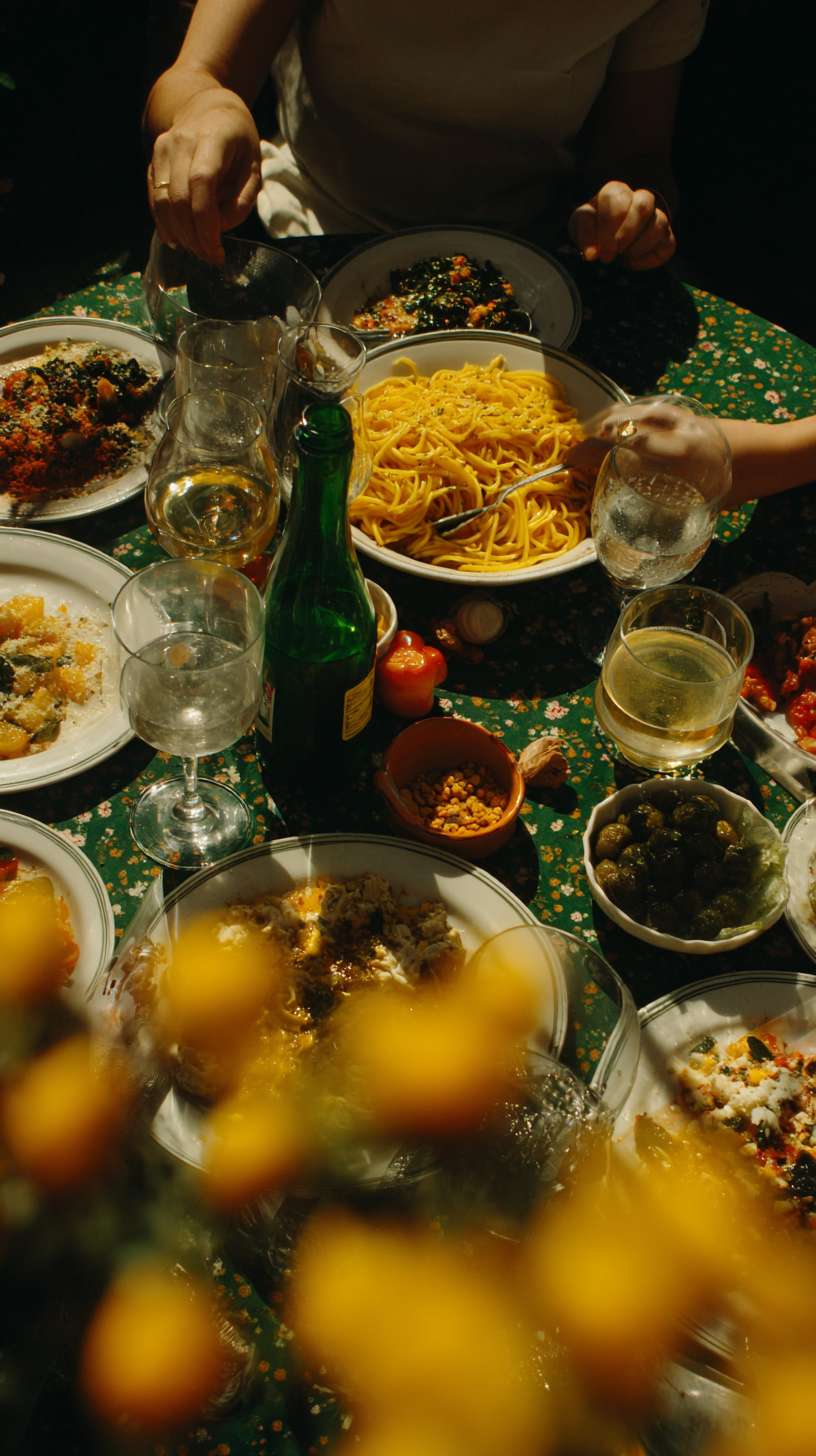 A table set with various dishes including spaghetti, salads, bowls of olives, wine glasses, and bottles, with a person serving food.
