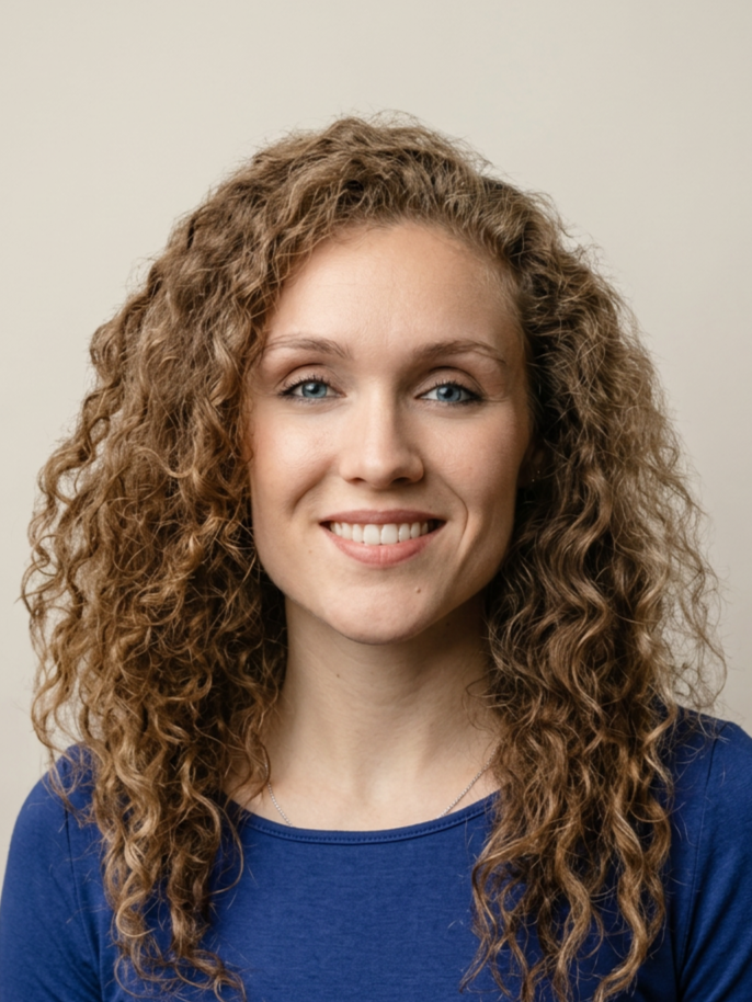 Portrait of a young woman with long curly red hair and blue eyes, smiling, wearing a blue shirt, against a plain beige background.