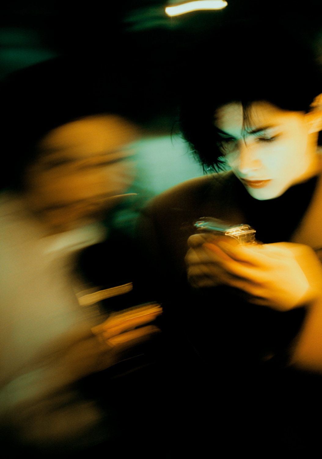 A woman with short black hair looking down at her phone in a dimly lit environment, with another person blurred in motion nearby.