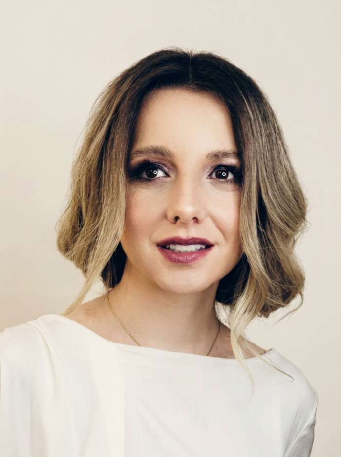 Portrait of a woman with shoulder-length wavy hair, wearing a white top, and subtle makeup, looking at the camera against a plain background.