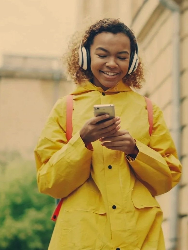 A young woman in a yellow raincoat with red straps is smiling and looking at her phone while wearing headphones outdoors.
