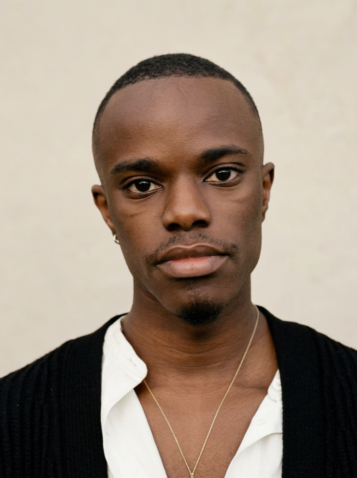 Close-up portrait of a young African American man with short hair, a small mustache, and a goatee, wearing a white shirt, black cardigan, and a silver chain necklace against a plain background.