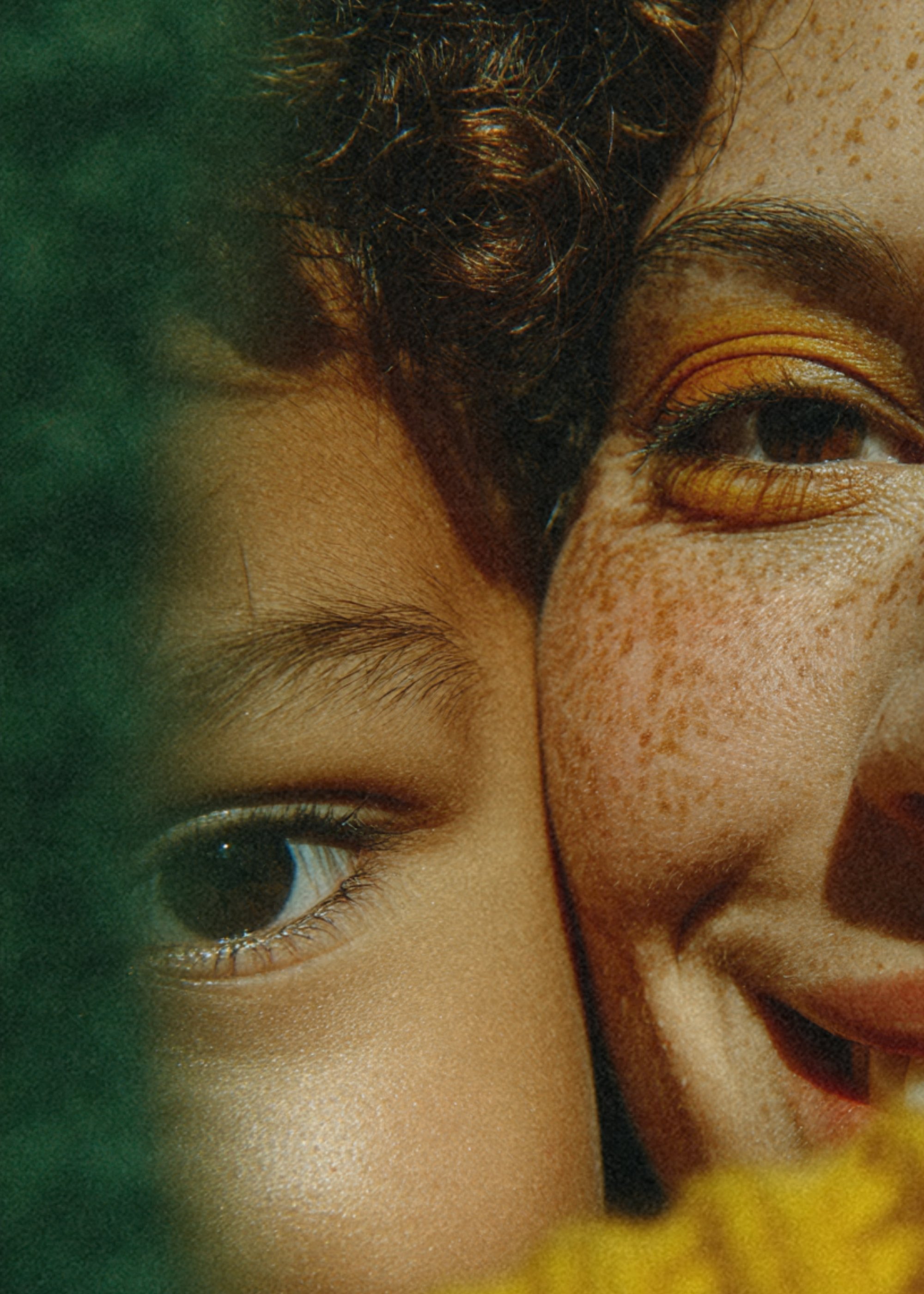 Close-up of a person's eye, eyebrow, and part of their face with freckles, showing dark curly hair and a background with blurred green and yellow elements.