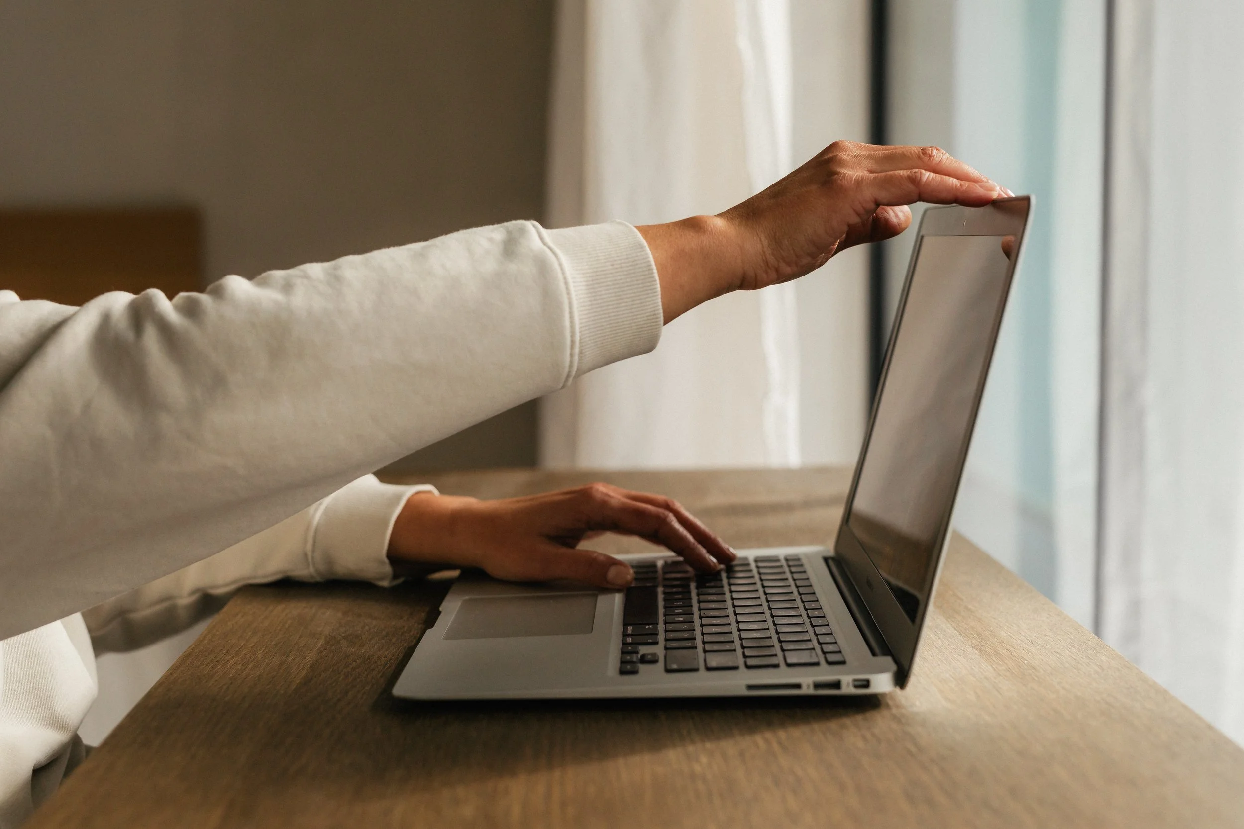 A person wearing a long sleeve shirt is using a laptop computer on a wooden desk near a window with light-colored curtains.