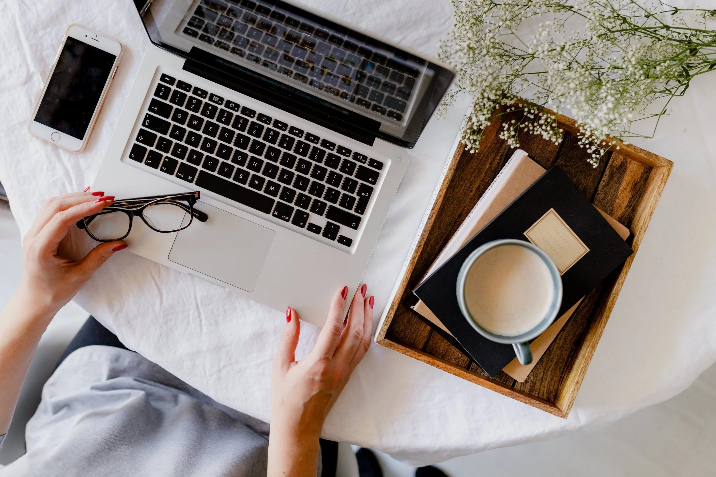 A person with red nails holding black glasses over a silver laptop with an open keyboard, on a white table with a smartphone nearby, a wooden tray with a coffee cup, notebooks, and a vase with white flowers.
