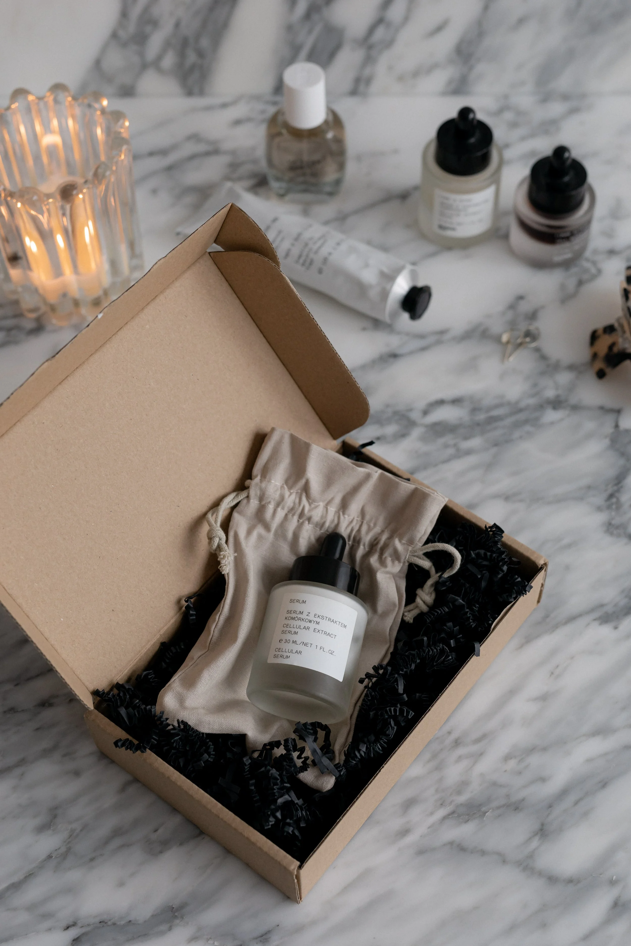 An open cardboard box on a marble surface containing a white serum bottle, black shredded paper, and beige tissue paper. Additional skincare or cosmetic bottles are placed on the marble surface outside the box, including a small clear bottle with a black cap, two dropper bottles, and a tube.