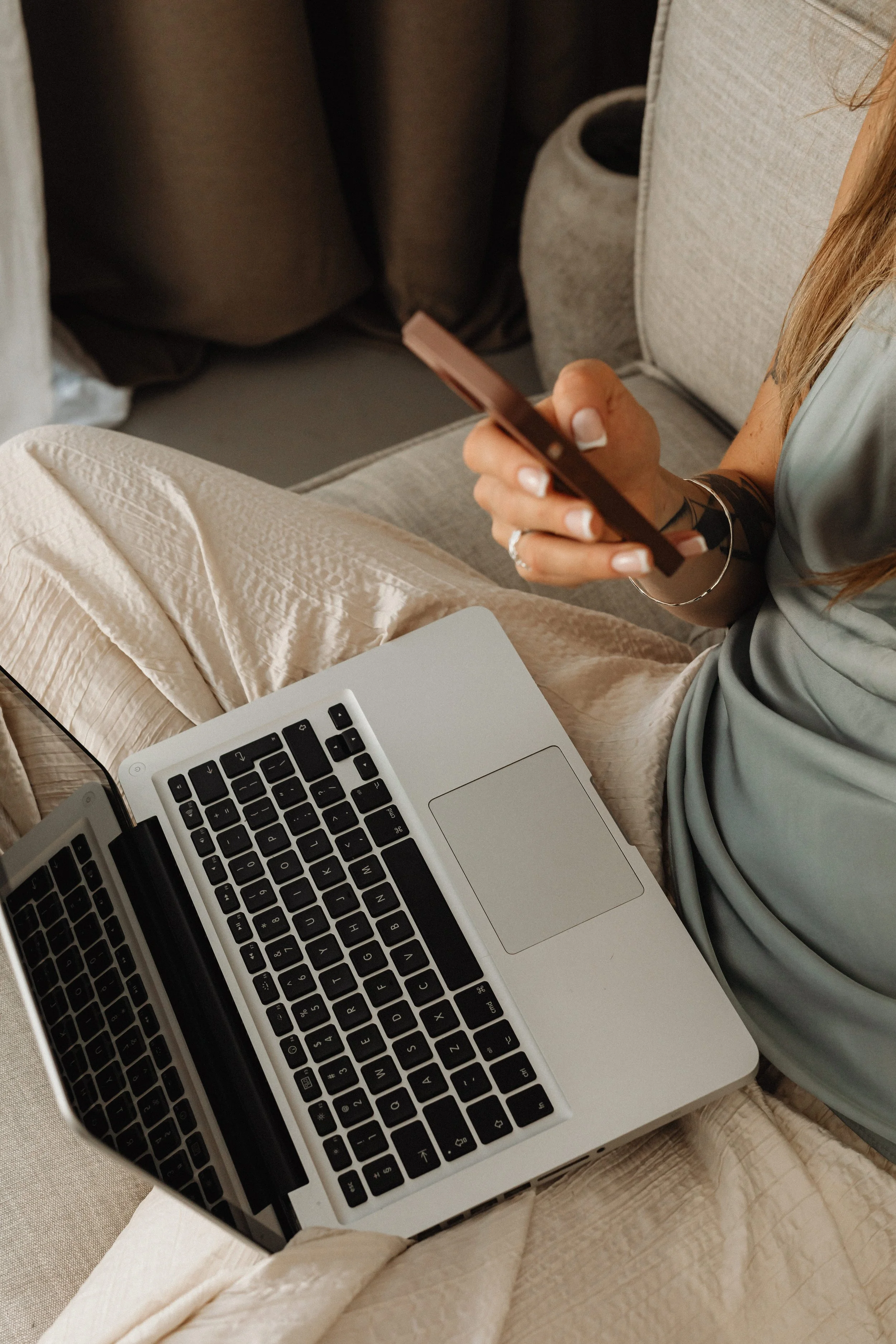 A person sitting on a beige couch, using a smartphone with a laptop open on their lap, wearing light-colored pants and a gray or shiny sleeveless top.