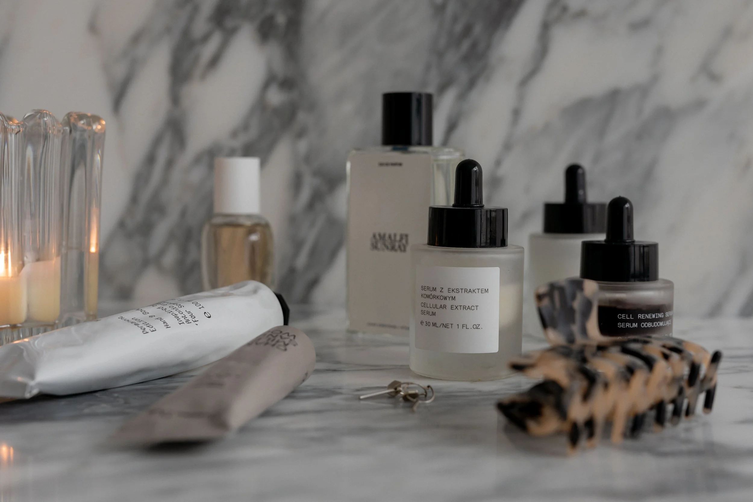 Collection of skincare and cosmetic bottles and tubes on a marble countertop, including serum dropper bottles and a tube with a white label.