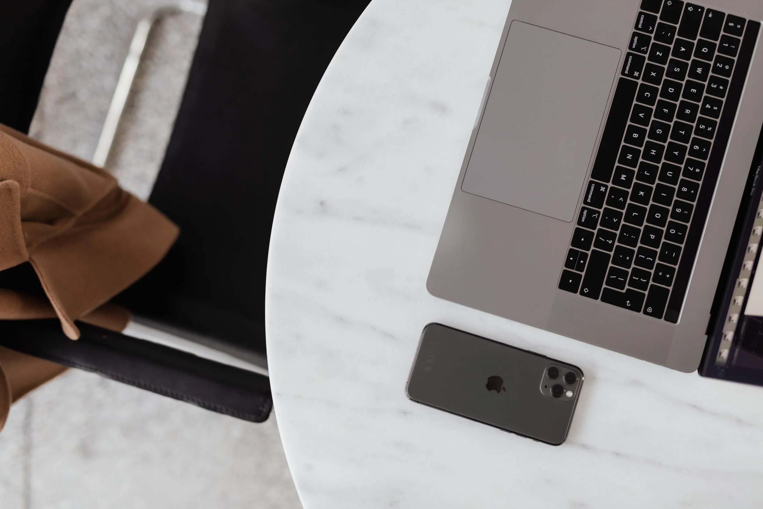 A close-up view of a silver MacBook laptop open on a white marble table, with a silver iPhone placed next to it. Part of a person's brown coat and blue jeans and black chair are also visible.