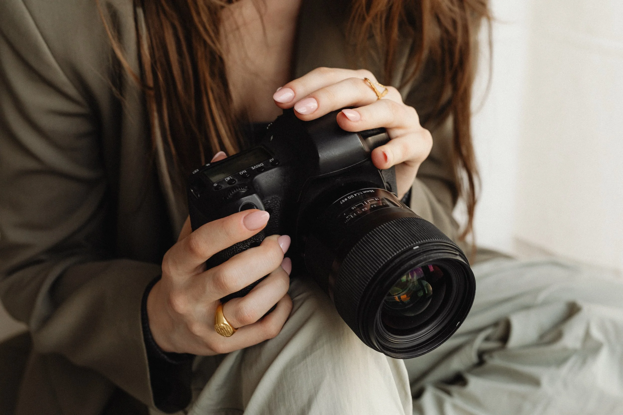 Close-up of a woman's hands holding a camera, with the focus on her fingers and rings, and her long hair visible in the background.