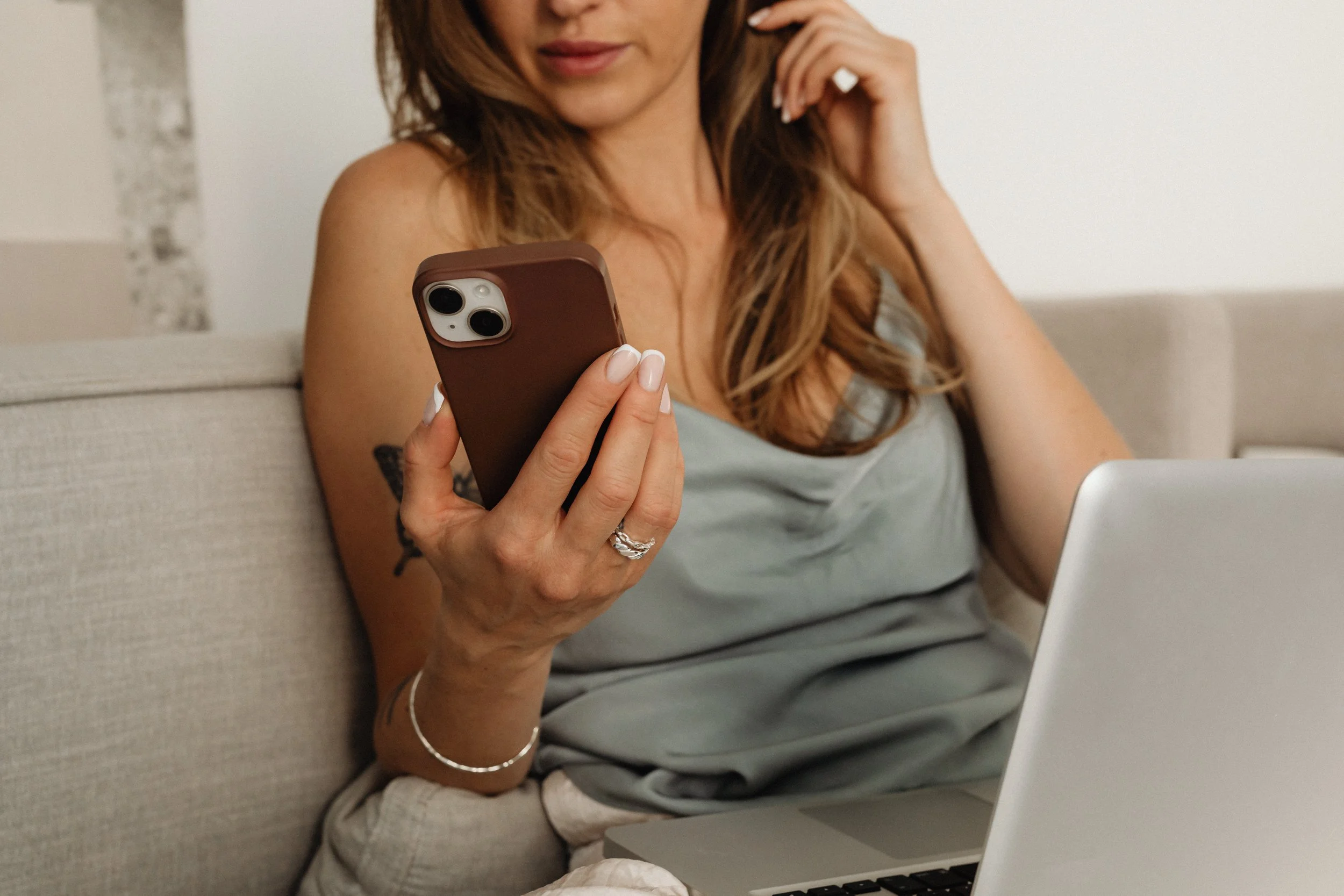 A woman sitting on a beige couch, looking at her smartphone. She has long, wavy hair, and is wearing a gray sleeveless top, a bracelet, and a ring. A laptop is on her lap, and the background shows a white wall with a decorative element.