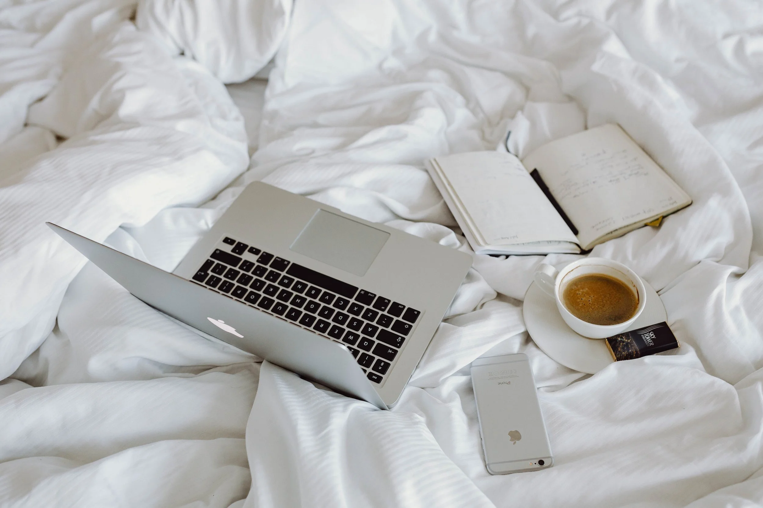A laptop, open notebooks, a cup of coffee, a smartphone, and a book are placed on a white bed with rumpled sheets.