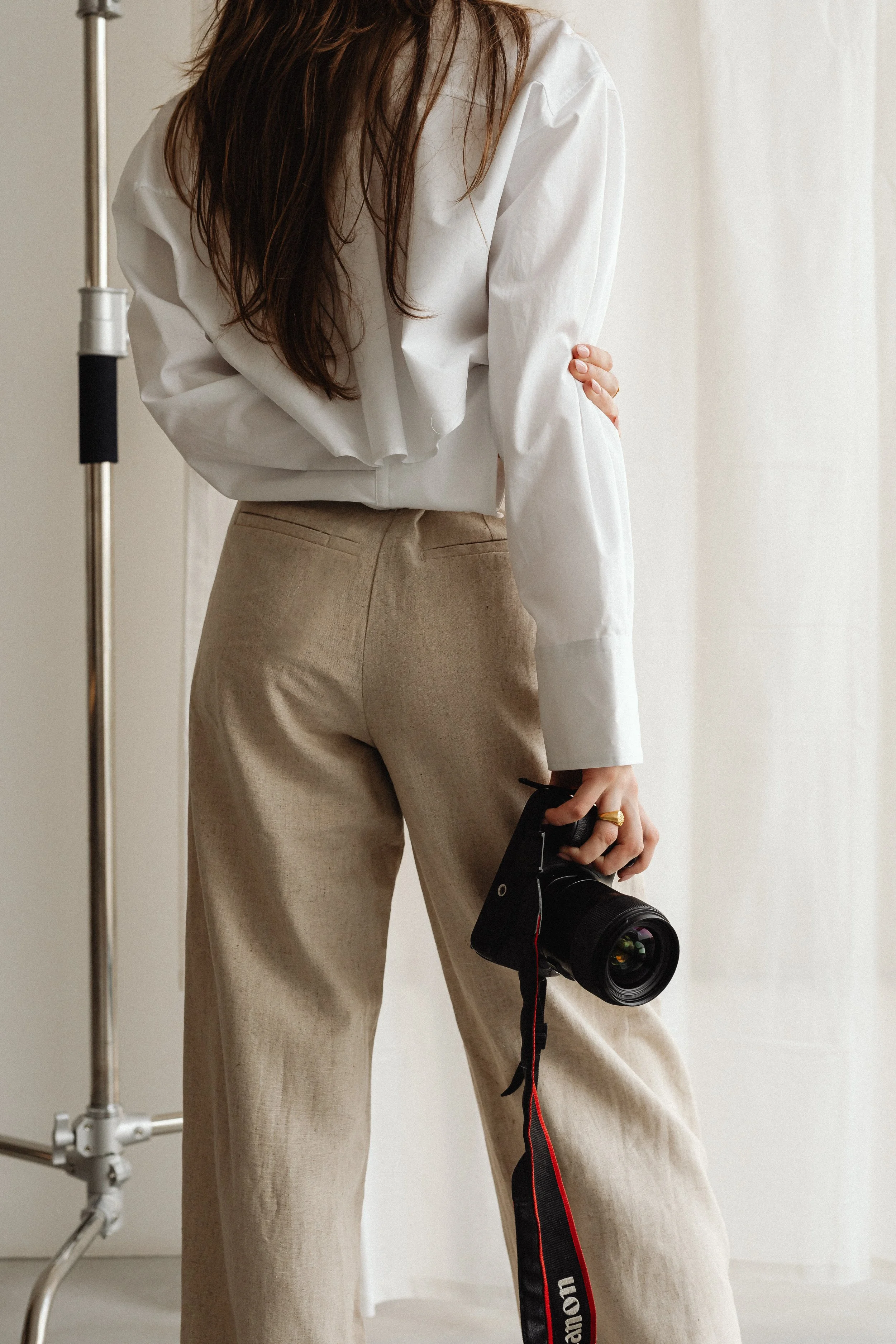 A woman in beige pants and a white shirt holding a professional camera with a large lens, standing indoors near a white wall and a light stand.