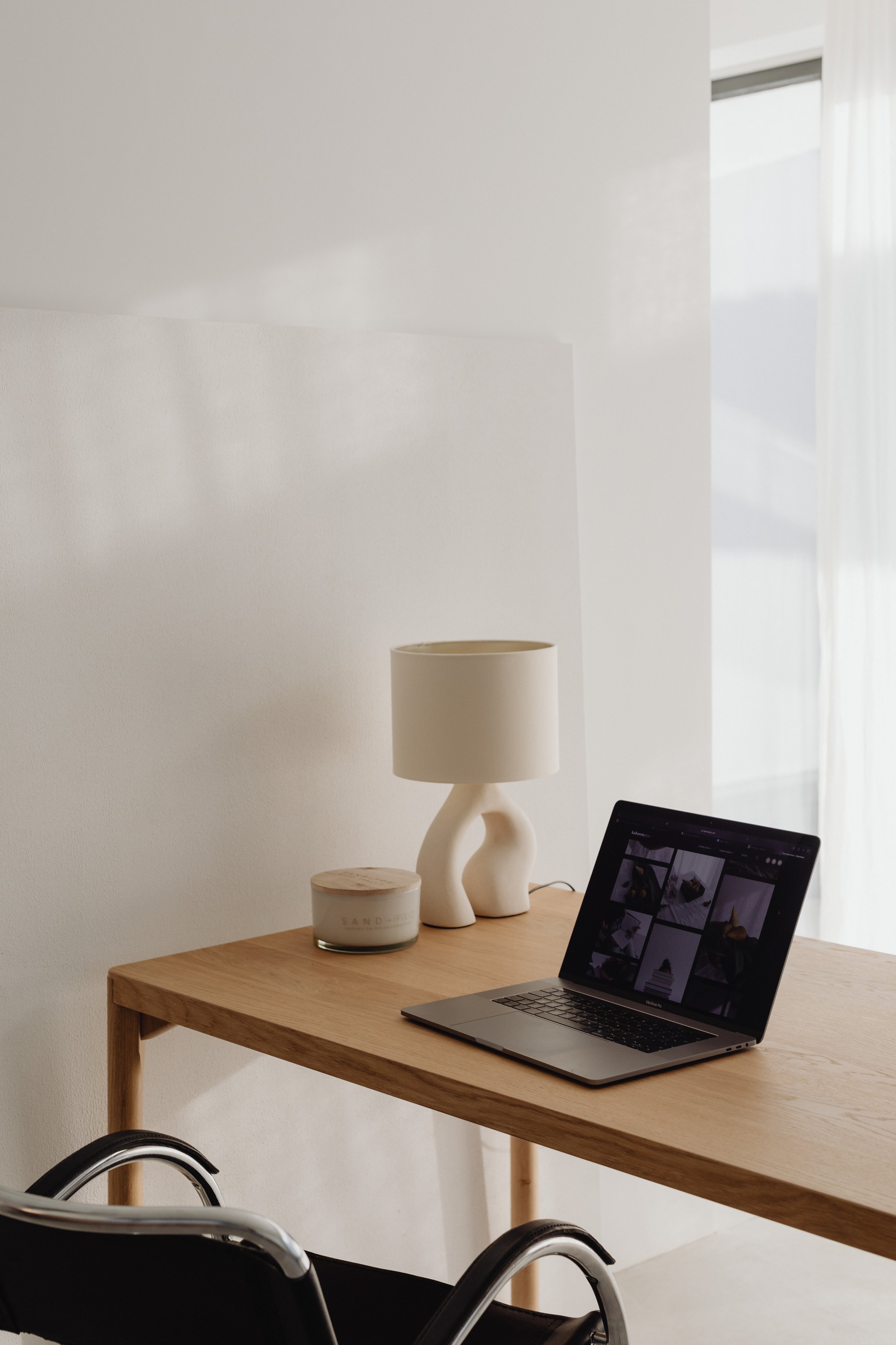 A wooden table holding a MacBook laptop with a grid of images on screen, a white ceramic lamp, a candle in a glass container, and a decorative abstract white sculpture. There are two black chairs with metal frames in front of the table, and in the background, a wall, a window with white curtains, and a wall-mounted lamp are visible.
