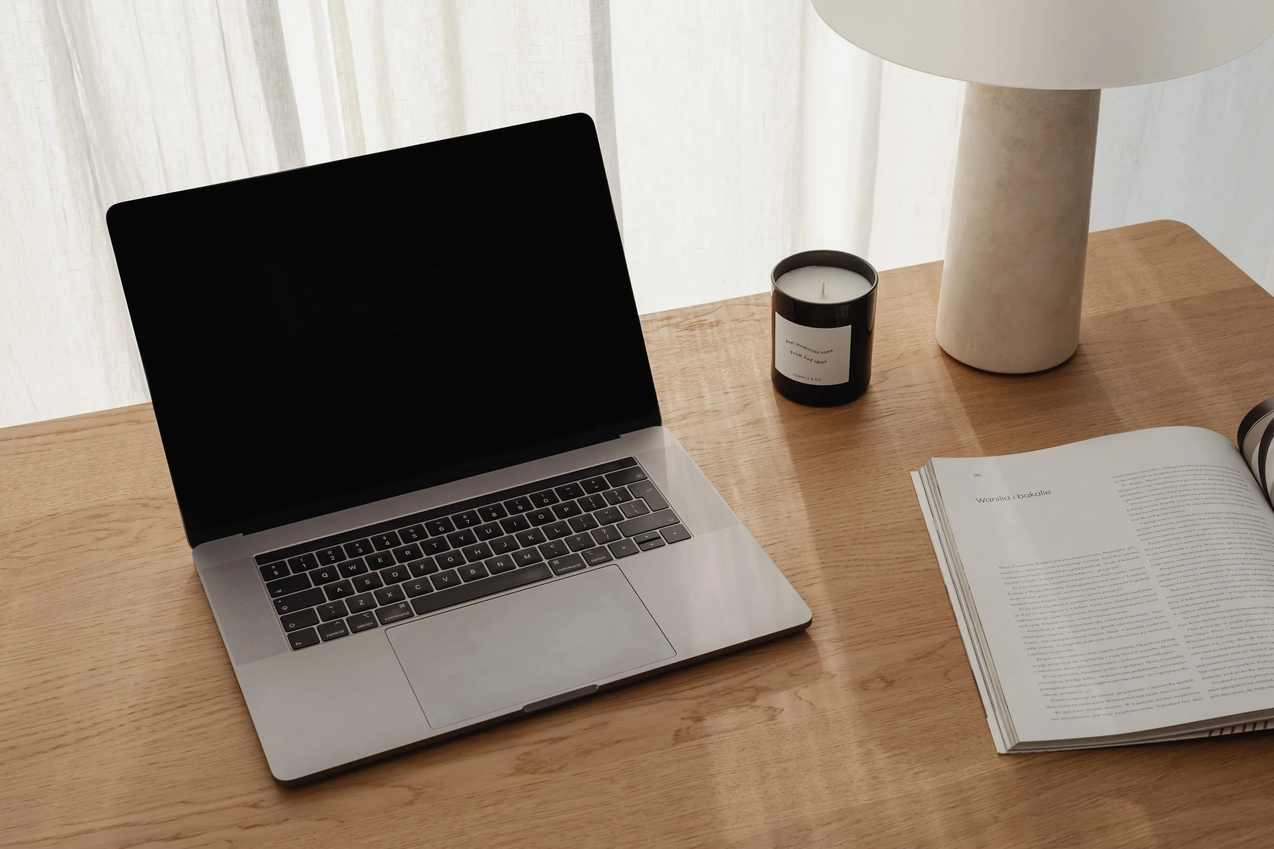 A wooden desk with an open laptop, a black candle, an off-white table lamp, and an open book with text.