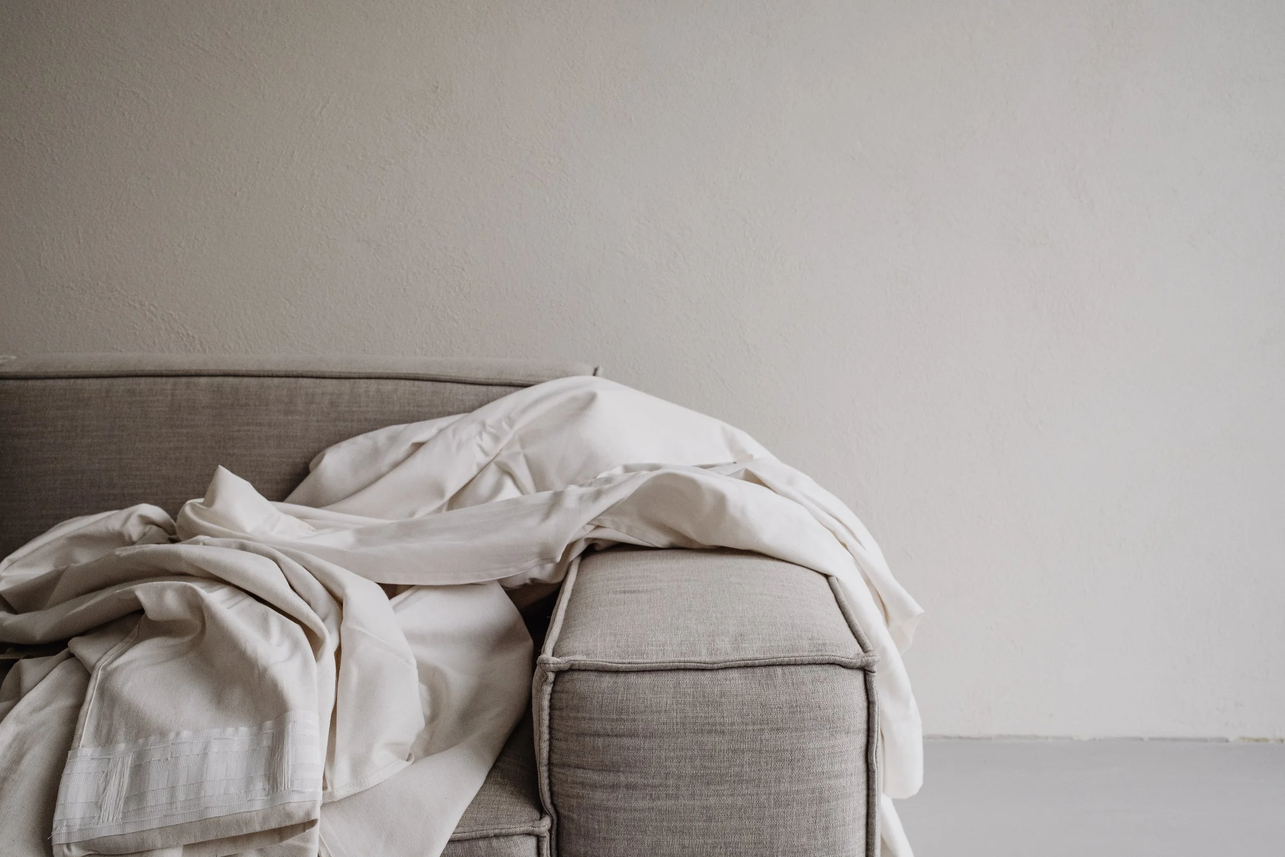 Disheveled white bed with crumpled sheets and a gray upholstered headboard against a plain beige wall.