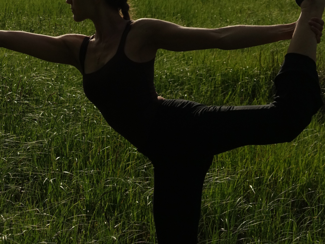 Eine Person macht Yoga im Stehen in einem grünen Feld, hält den Fuß mit der Hand und dehnt sich.
