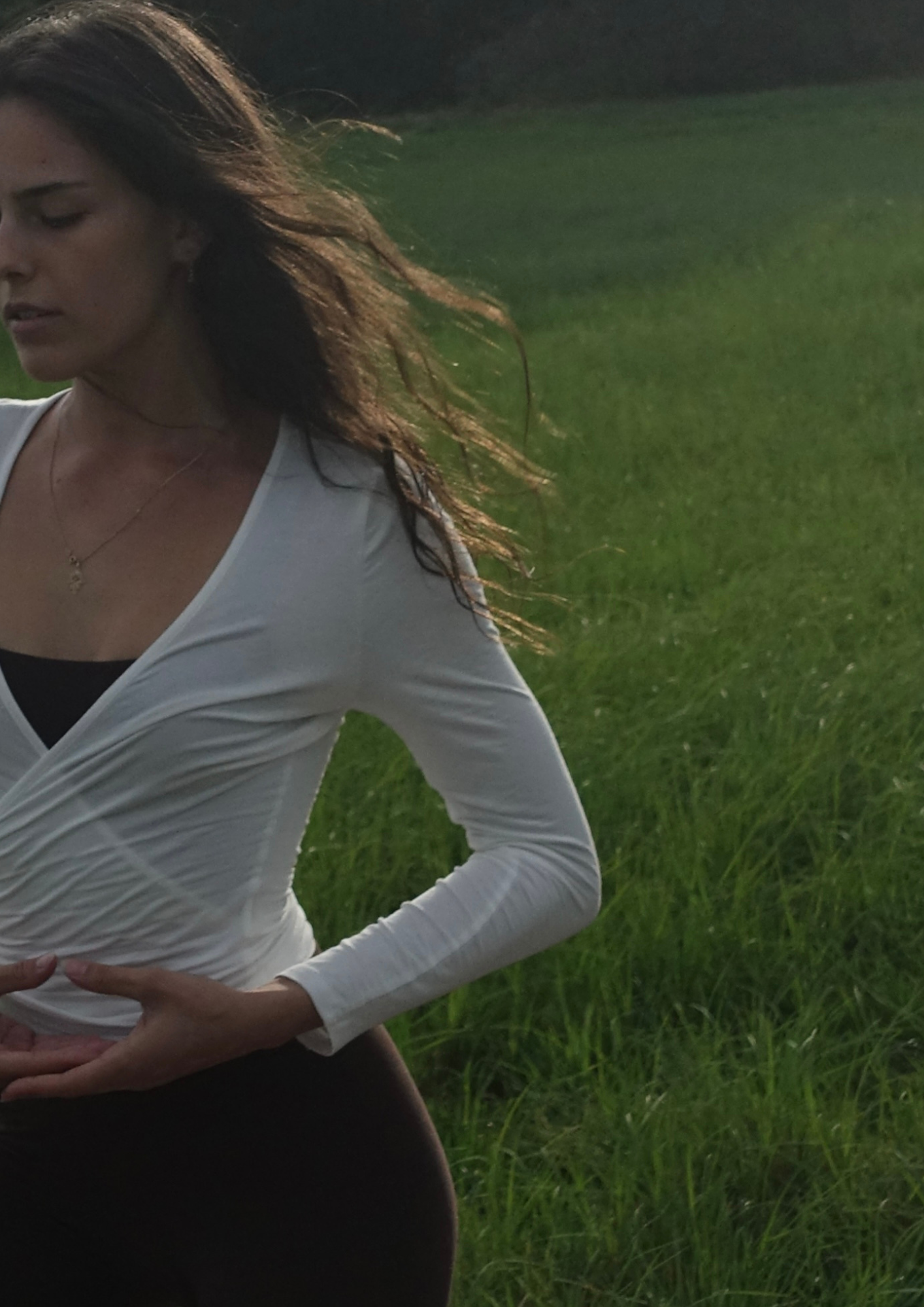 Eine junge Frau in weißem Oberteil, die an einem grünen Feld steht, bei Sonnenuntergang oder -aufgang, mit langen, gewellten braunen Haaren, die im Wind wehen.