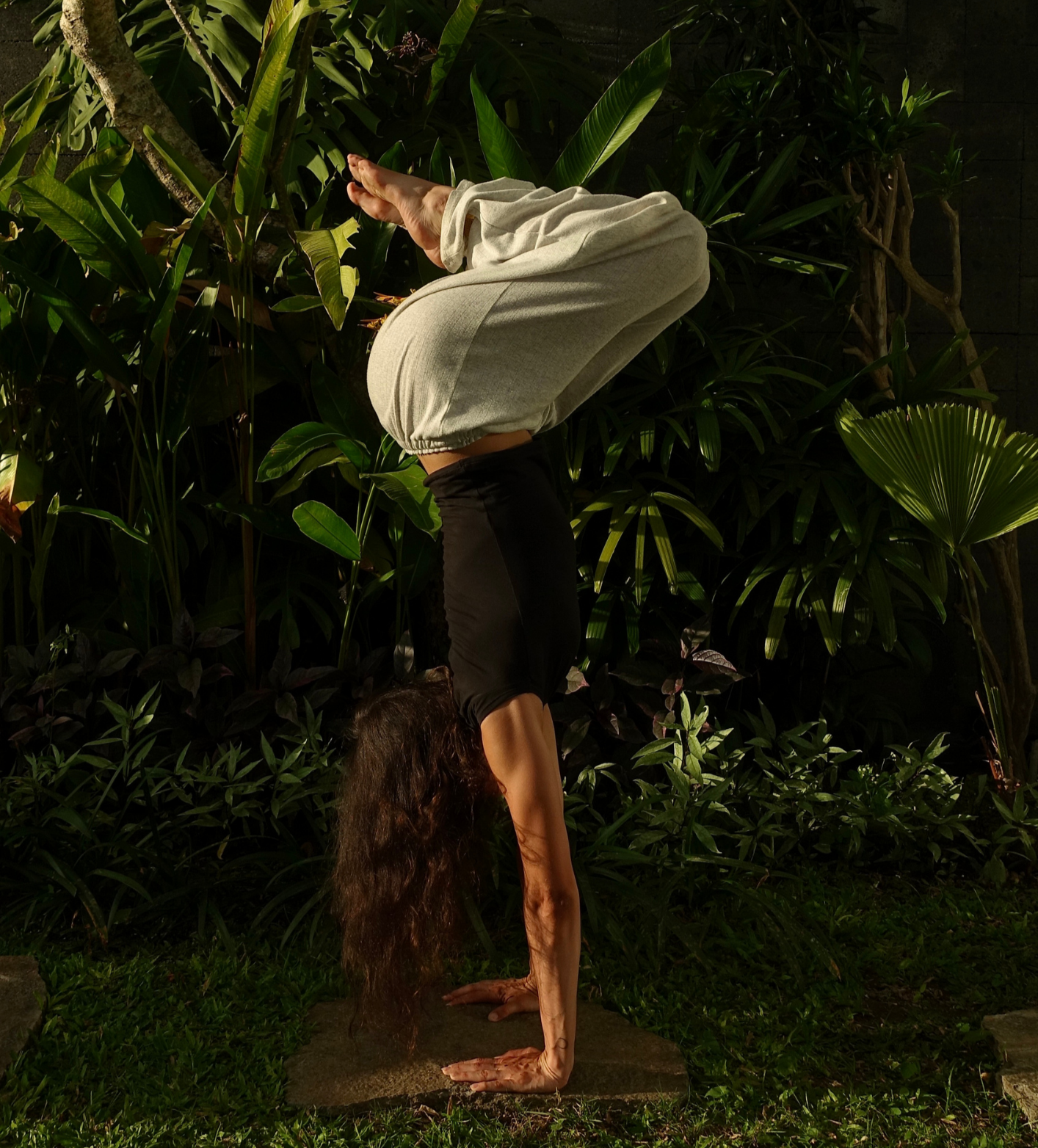 Frau übt den Handstand in einem Garten mit vielen grünen Pflanzen.