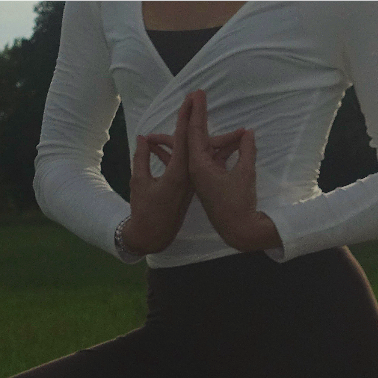 Person, die in einer Yoga-Pose steht, mit den Händen in der Mudra-Position, trägt ein weißes langärmeliges Top und schwarze Kleidung, im Freien bei Dämmerlicht.
