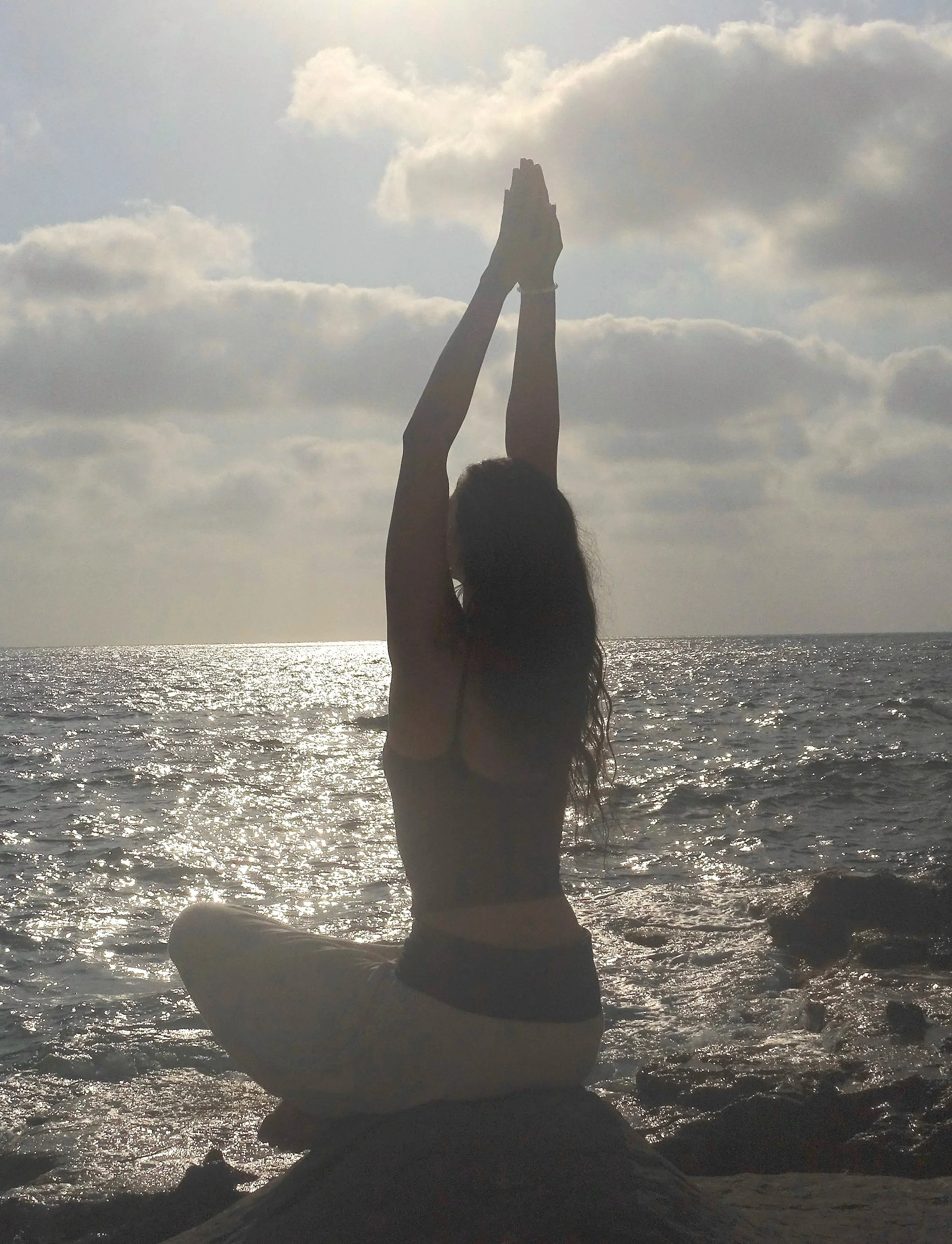 Frau übt Yoga am Strand mit gestreckten Armen und verschränkten Händen, Blick Richtung Himmel, im Hintergrund das Meer und bewölkter Himmel.