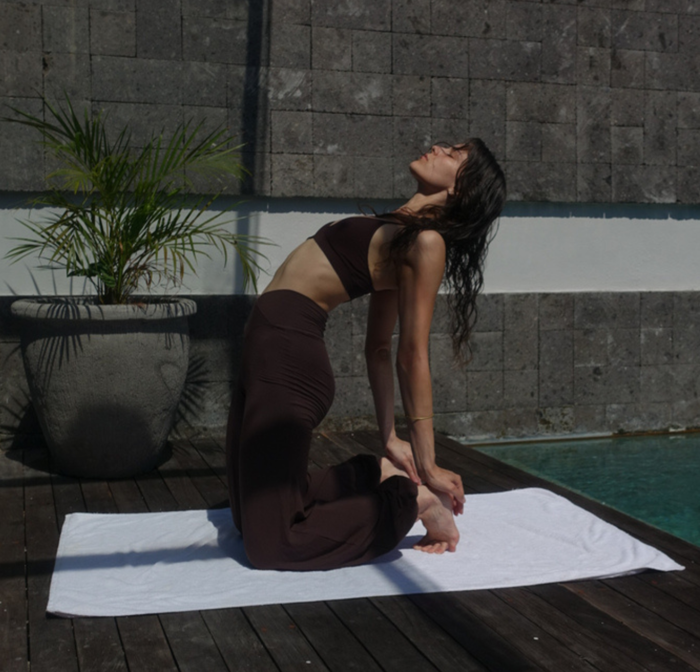 Frau in Yoga-Haltung auf einer weißen Matte neben einem Pool im Freien, mit Ziegelmauer im Hintergrund und einer großen Topfpflanze.