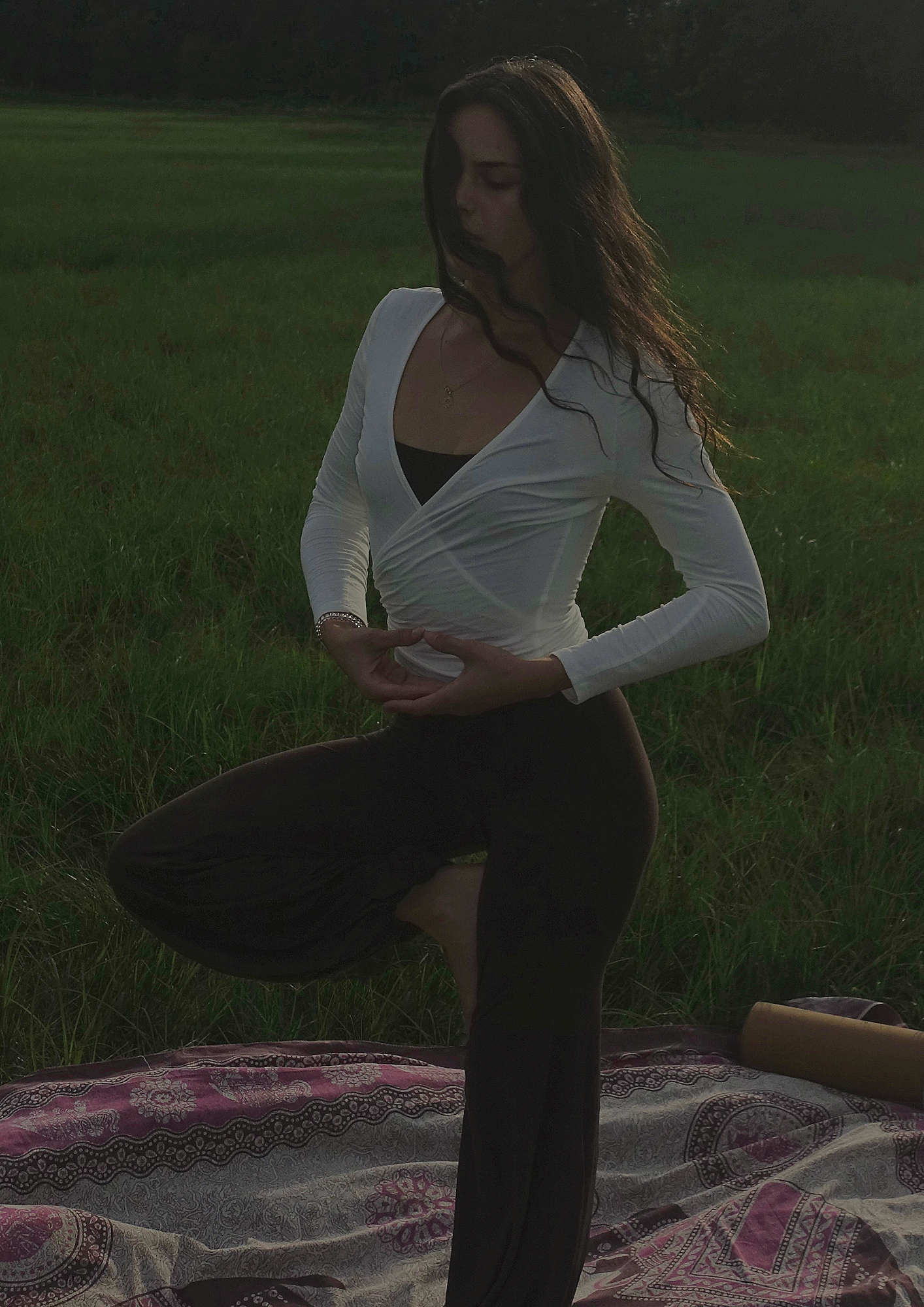 Eine Frau in einer Yoga-Pose im Freien bei Sonnenuntergang, auf einer Matte auf dem Boden, mit Grünflächen im Hintergrund.