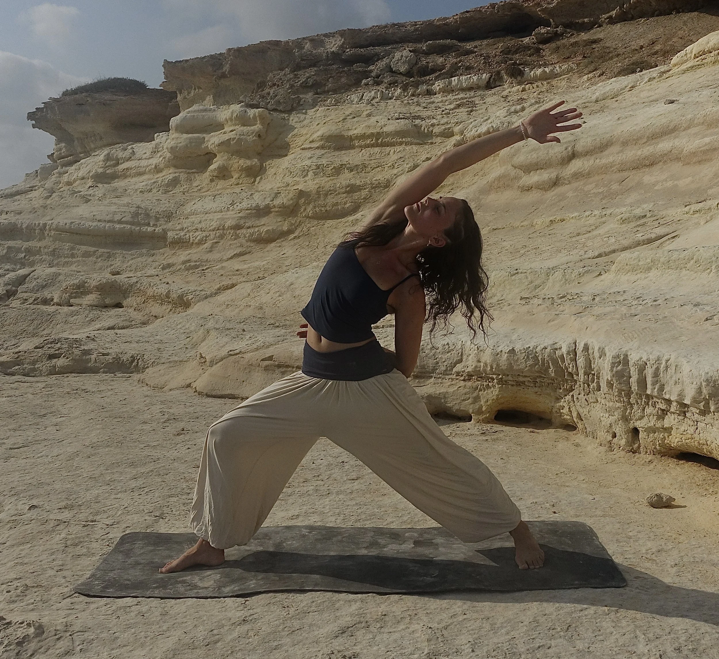 Eine Frau steht im Yoga-Tierkörper, auf einer Yoga-Matte auf einem Sandstrand, mit Felsen im Hintergrund, Sonnenlicht auf ihrem Gesicht, trägt eine schwarze Sportoberbekleidung, ist entspannt und konzentriert.