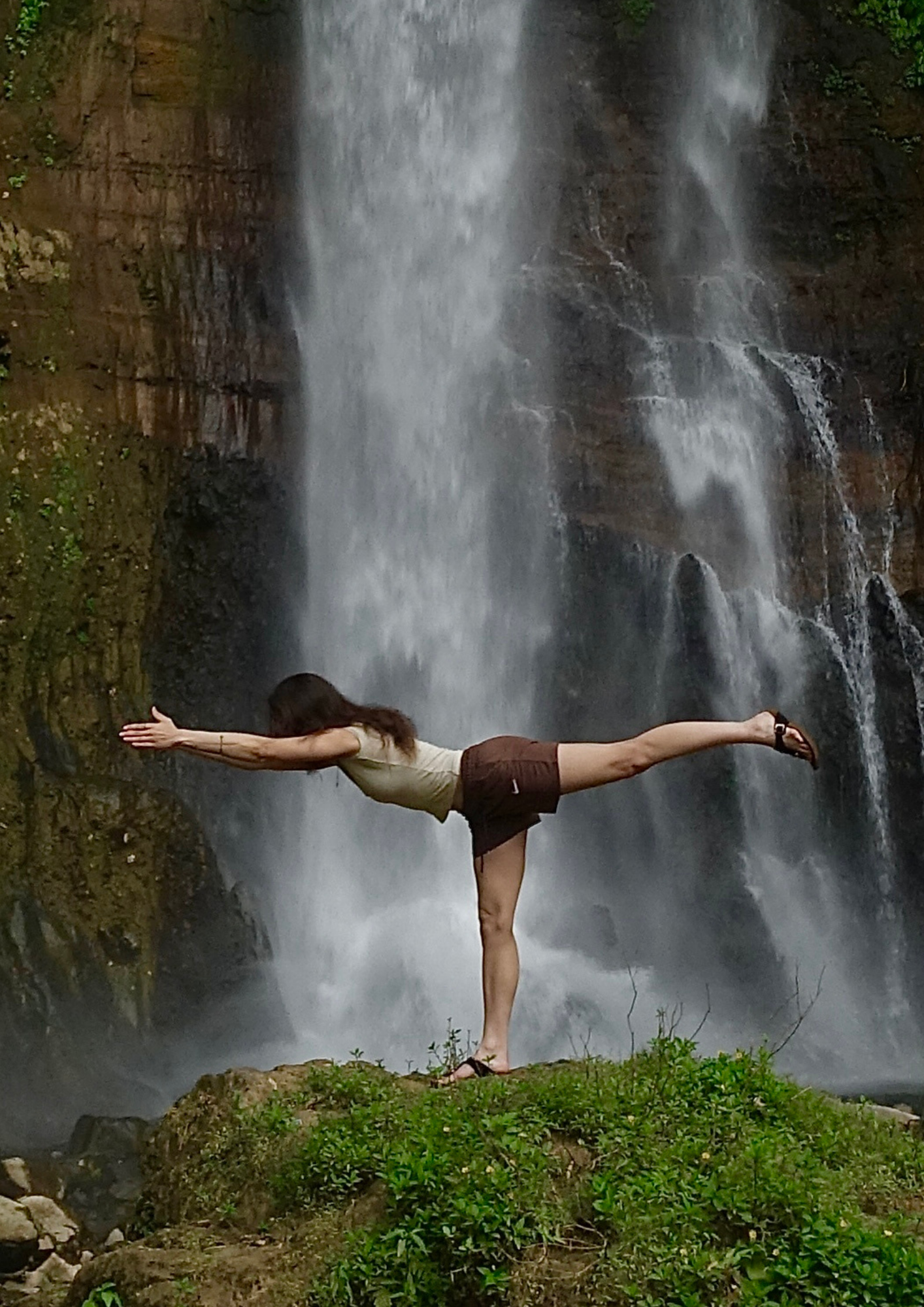 Eine Frau in Yoga-Pose vor einem Wasserfall, auf einem Felsen im Grünen.