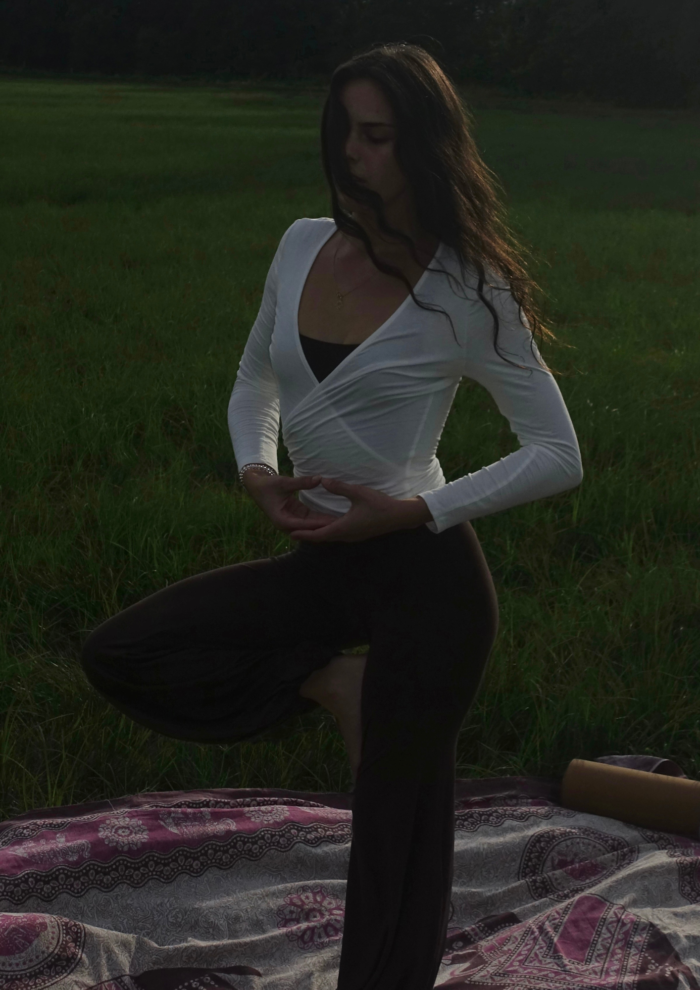 Frau praktiziert Yoga im Freien bei Sonnenuntergang auf einer Decke in einem grünen Feld.
