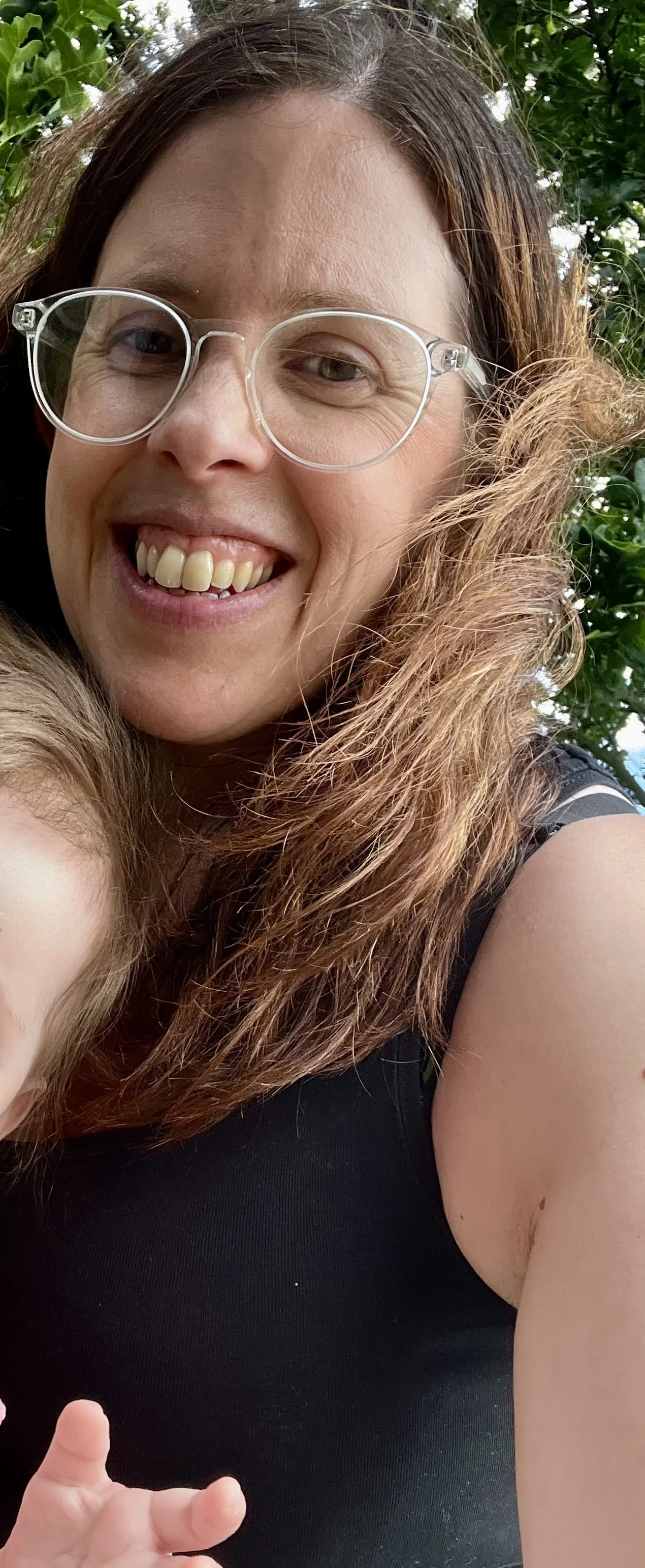 Close-up of a smiling woman with brown wavy hair and glasses, wearing a black sleeveless shirt, outdoors with green foliage in the background.