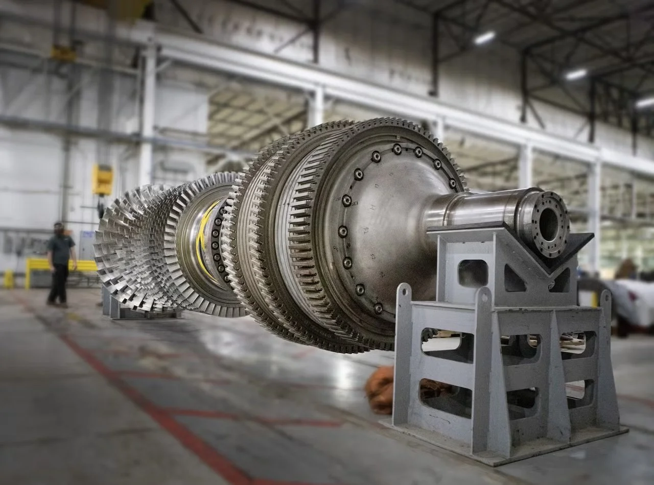 Large industrial turbine or engine part with multiple gear-like components, mounted on a stand inside a manufacturing workshop.