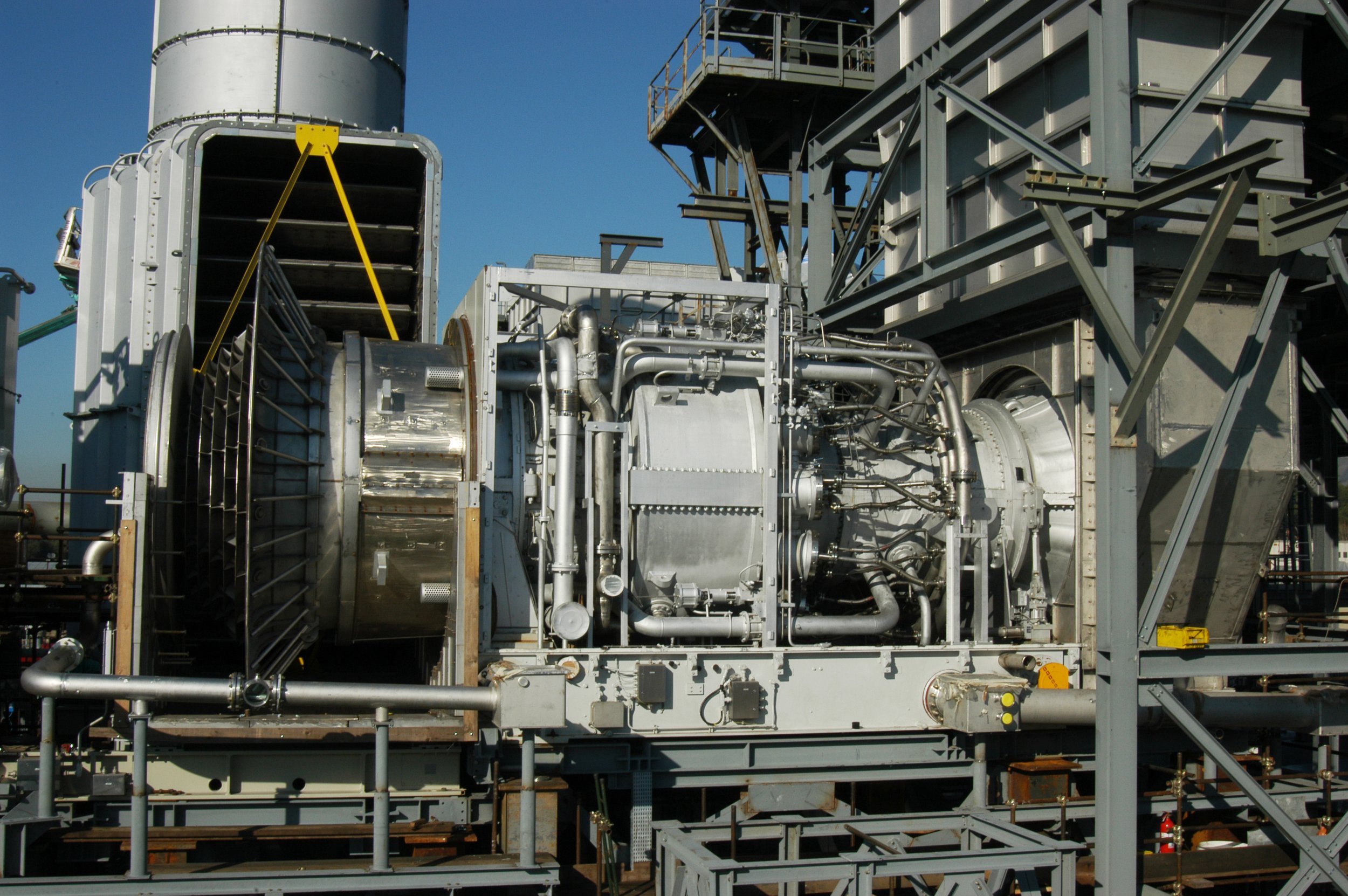 Industrial machinery with pipes, metal structures, and scaffolding against a clear blue sky.
