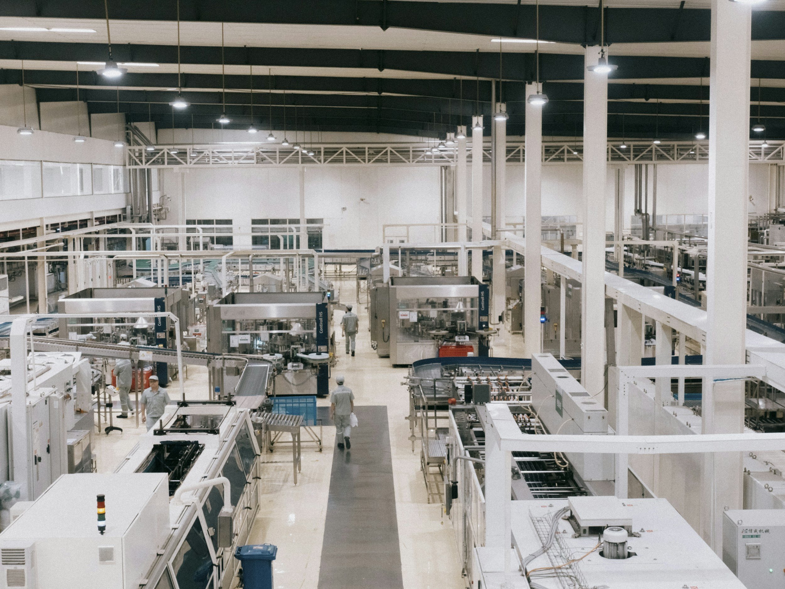 Interior of a large industrial or manufacturing facility with machinery, conveyor belts, and workers wearing white uniforms, including some with helmets. The space has high ceilings with hanging lights and structural beams.