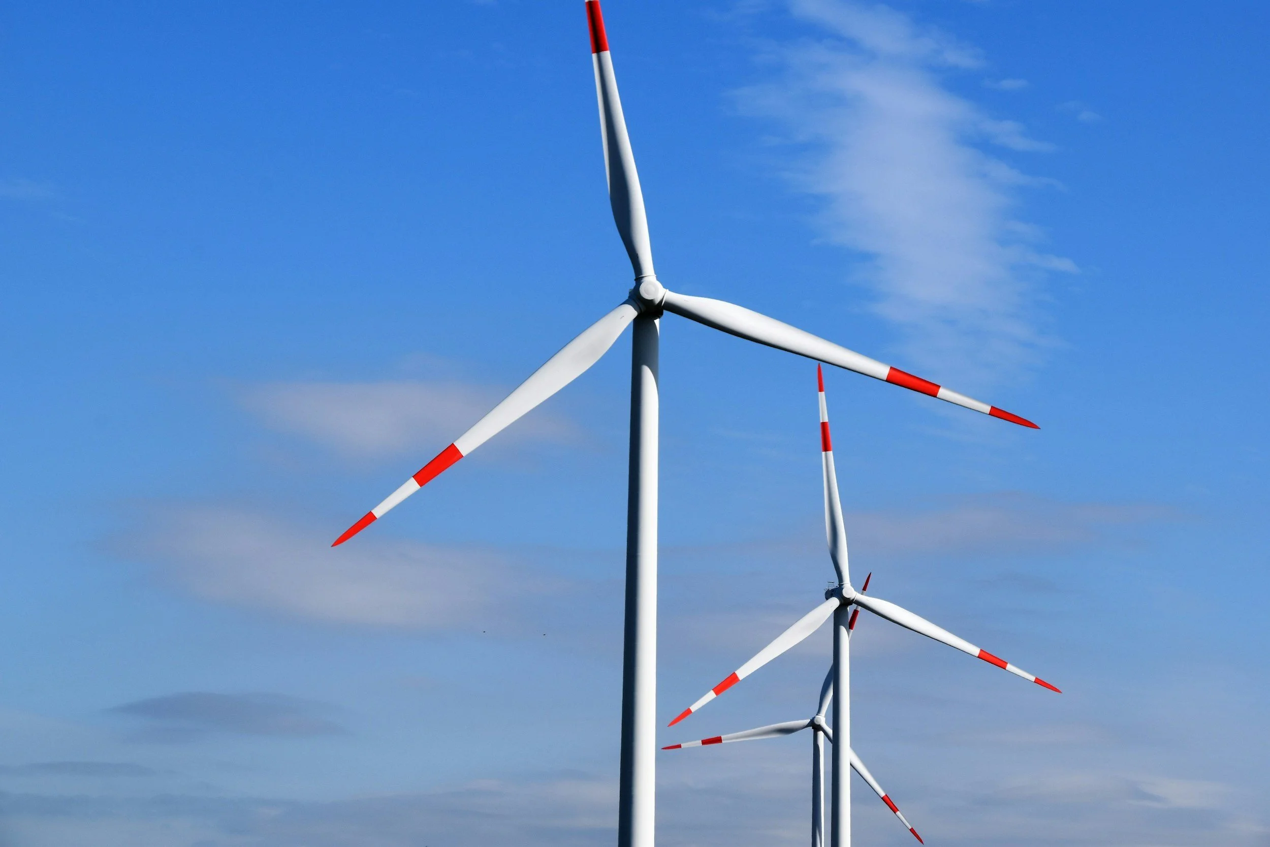 Multiple wind turbines with white blades and red tips against a blue sky with some clouds.