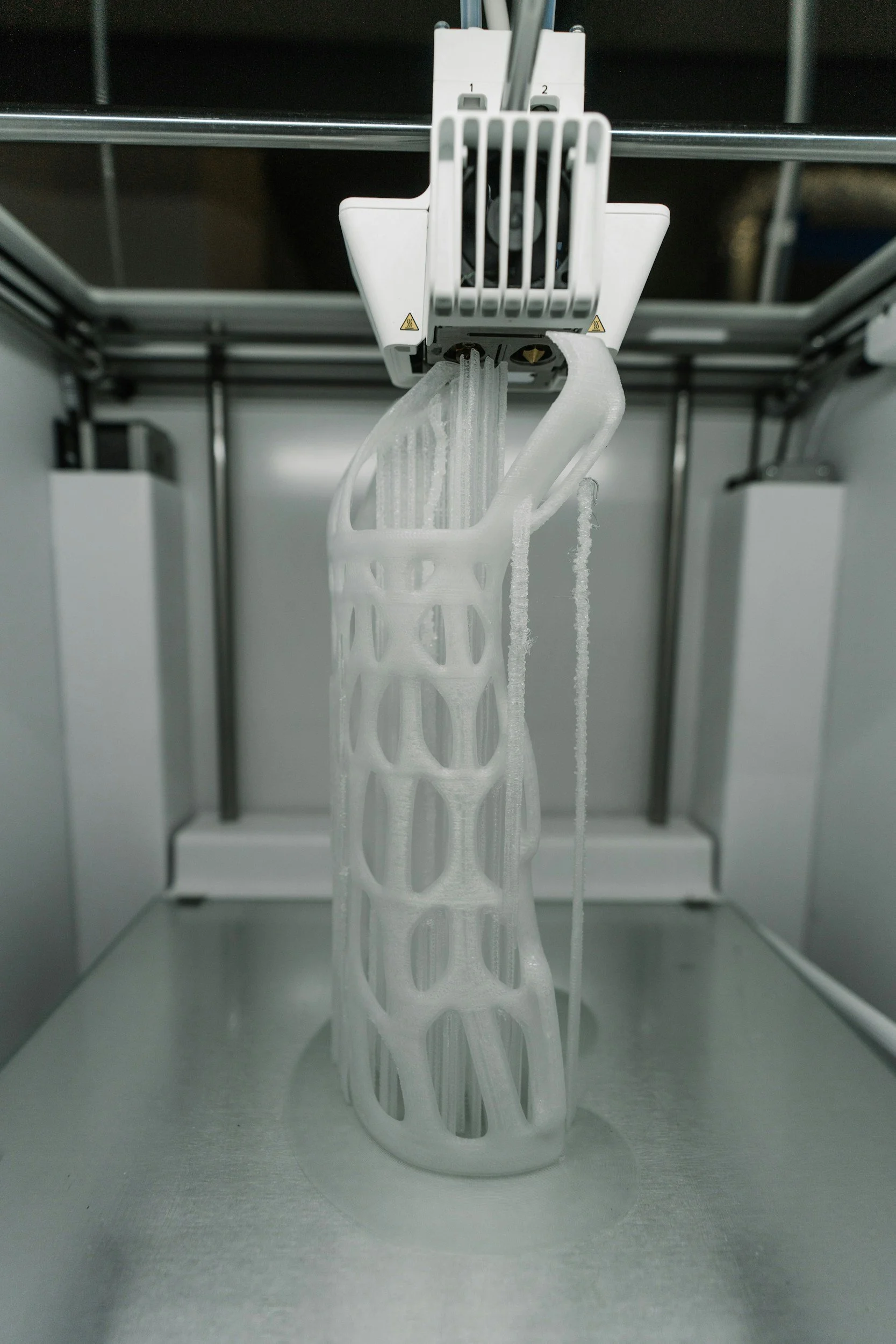A 3D printer nozzle is printing a lattice structure with white filament inside an enclosed 3D printing chamber.