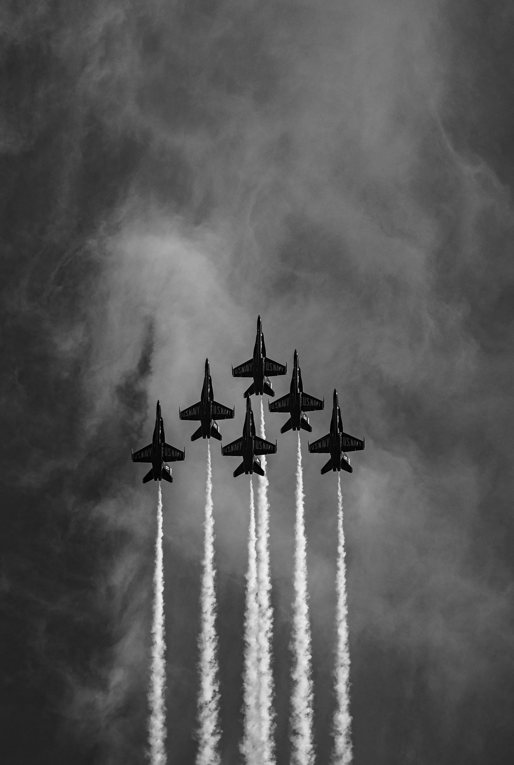 Six US Navy fighter jets flying in a tight formation, releasing smoke trails against a cloudy sky.