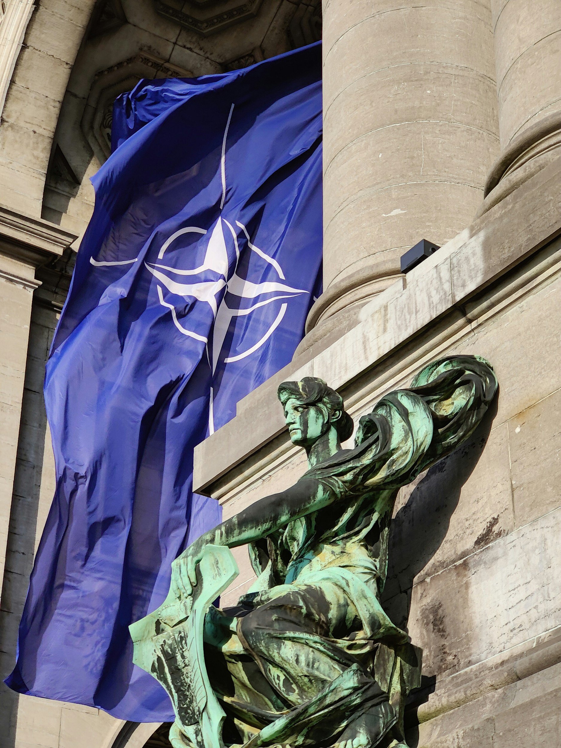A view of a building facade featuring large stone columns and an ornate bronze or copper sculpture of a woman holding a torch, with a blue flag displaying a white emblem hanging nearby.