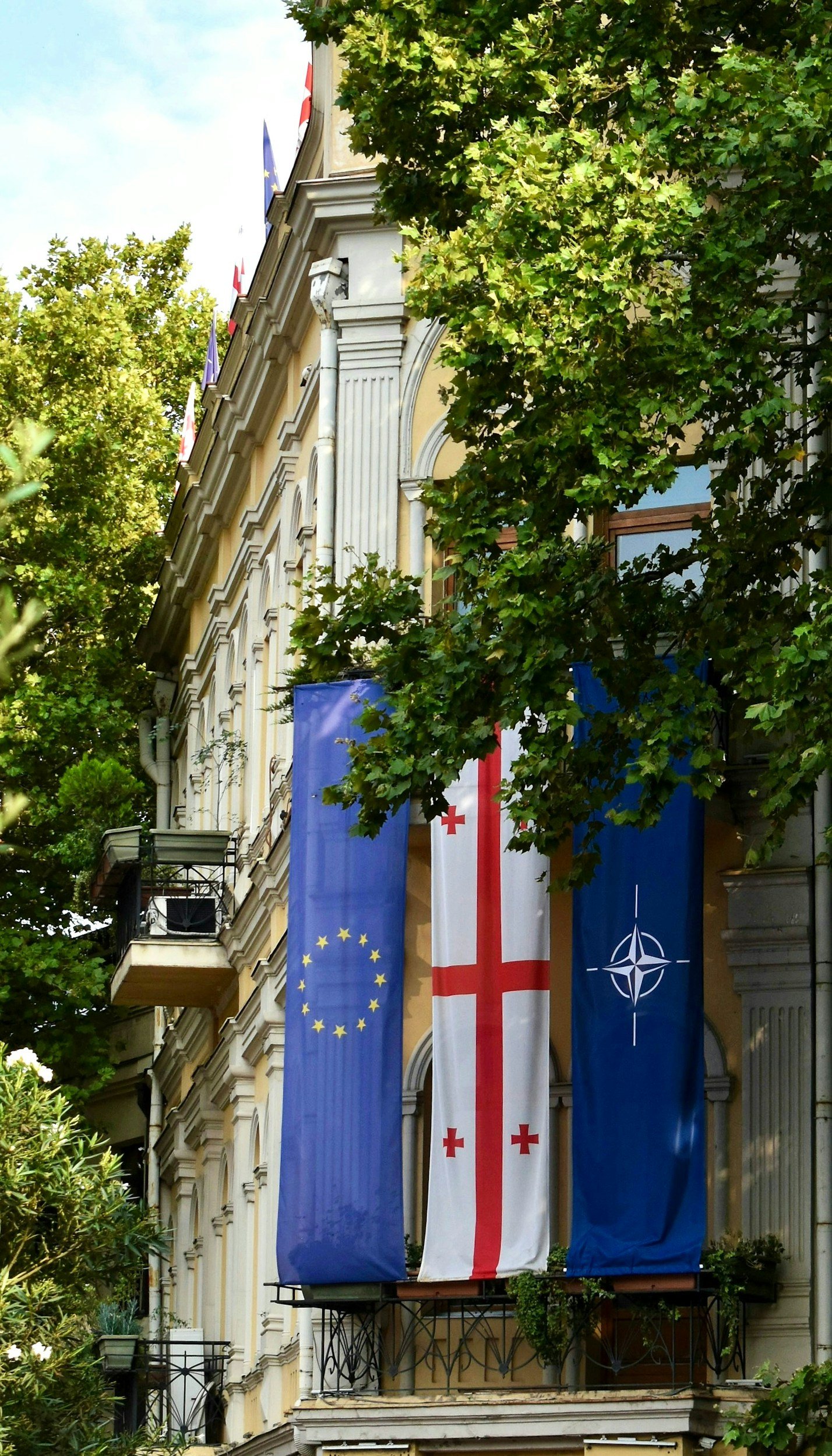 Flags hanging on the building with European Union, Georgia, and NATO flags, surrounded by green trees.