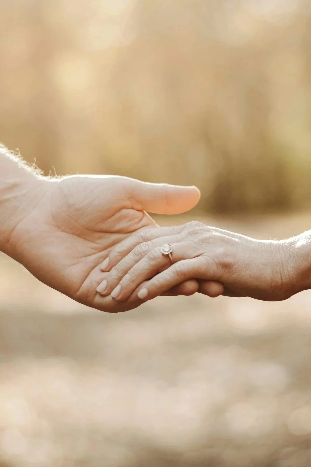Zwei Hände, die sich berühren, eine Hand mit einem Verlobungsring am Ringfinger. Helles, unscharfes Hintergrund.