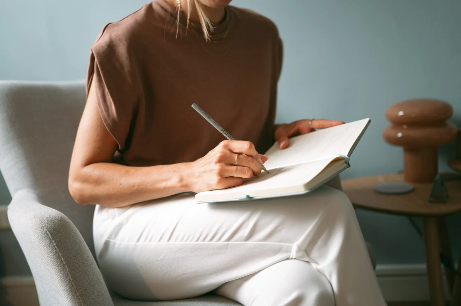 Frau sitzt in einem weißen Sessel und schreibt in ein Notizbuch mit einem grauen Stift. Im Hintergrund ist ein kleiner Tisch mit dekorativen Gegenständen zu sehen.