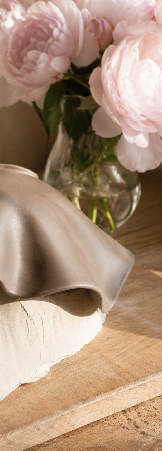 Nahaufnahme einer Belly Bowl, welche auf einem Gipsabdruck liegt. Die Schale ist organisch geschwungen mit welligen Rändern. Im Hintergrund stehen rosa Pfingstrosen in einer Glas-Vase.