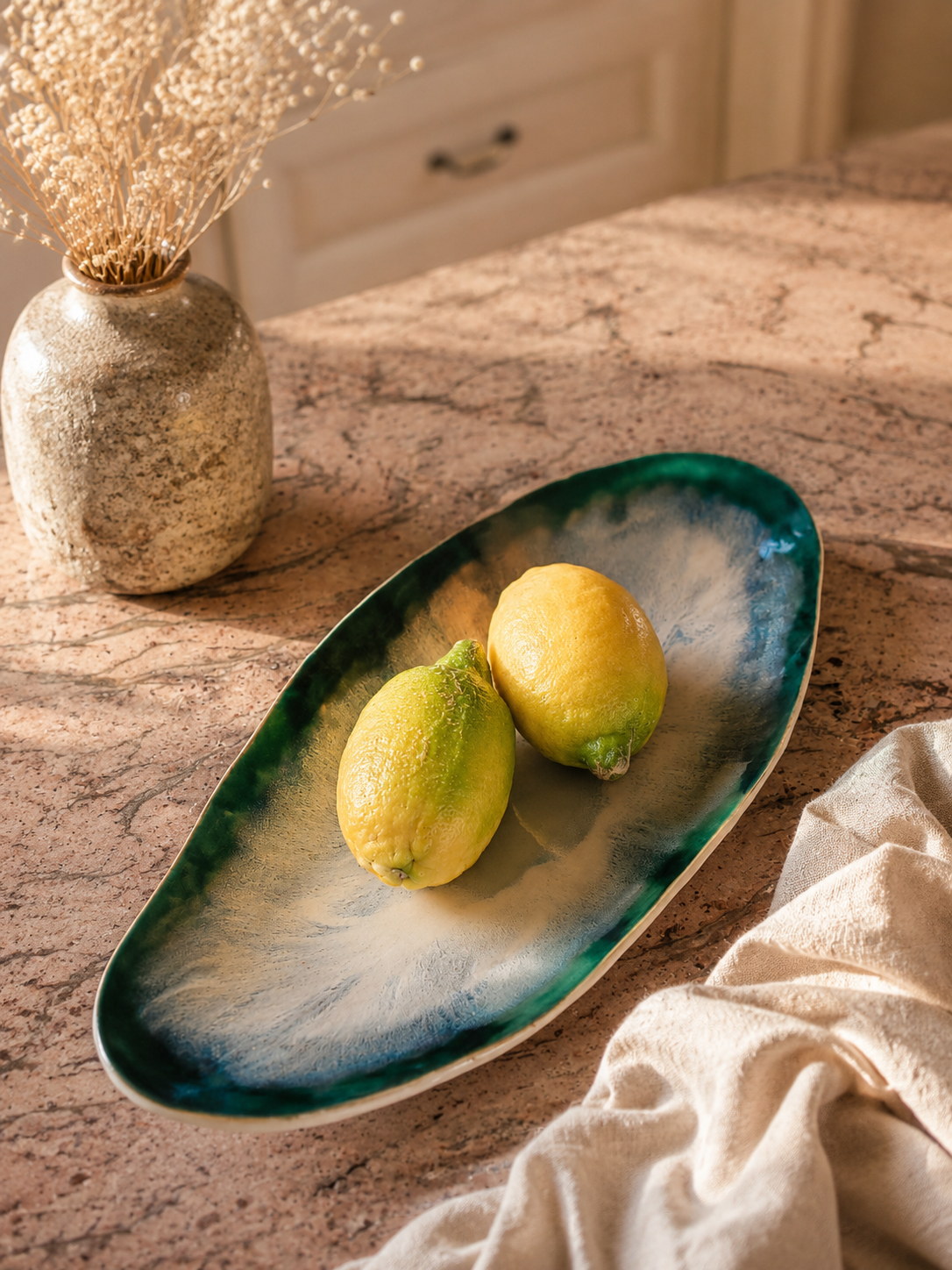 Fertige Belly Bowl in ovaler Form. Der Rand ist dunkelgrün glasiert und verläuft nach innen mit einer weißen Effekt-Glasur. Die Schale steht auf einer bräunlichen Marmorplatte und ist mit Zitronen arrangiert.