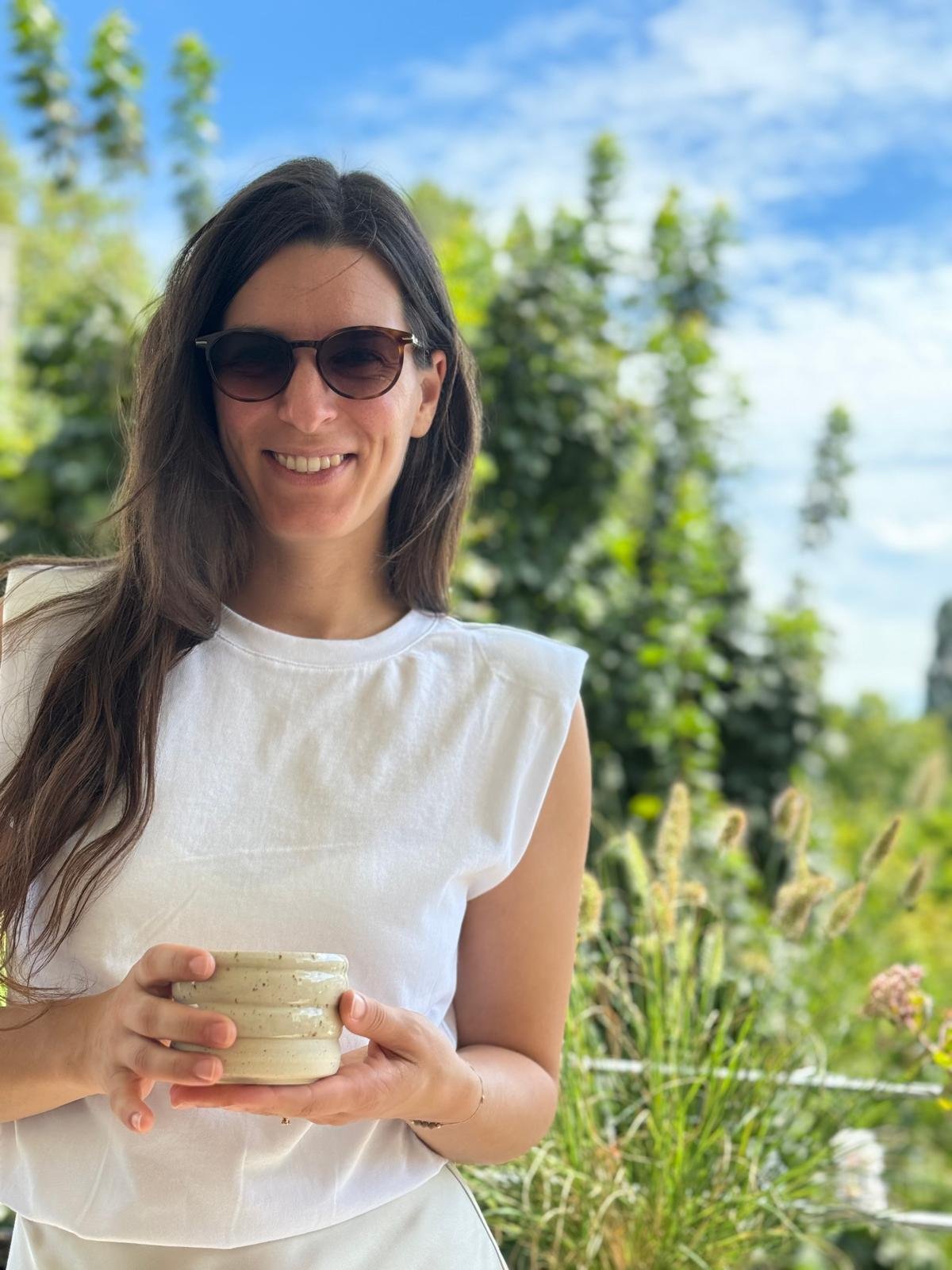 Saskia Lurker, Keramikerin und Herstellerin der Belly Bowls, mit braunen Haaren und einer handgemachten Keramiktasse in der Hand. Im Hintergrund sind viele Bäume und Natur erkennbar.